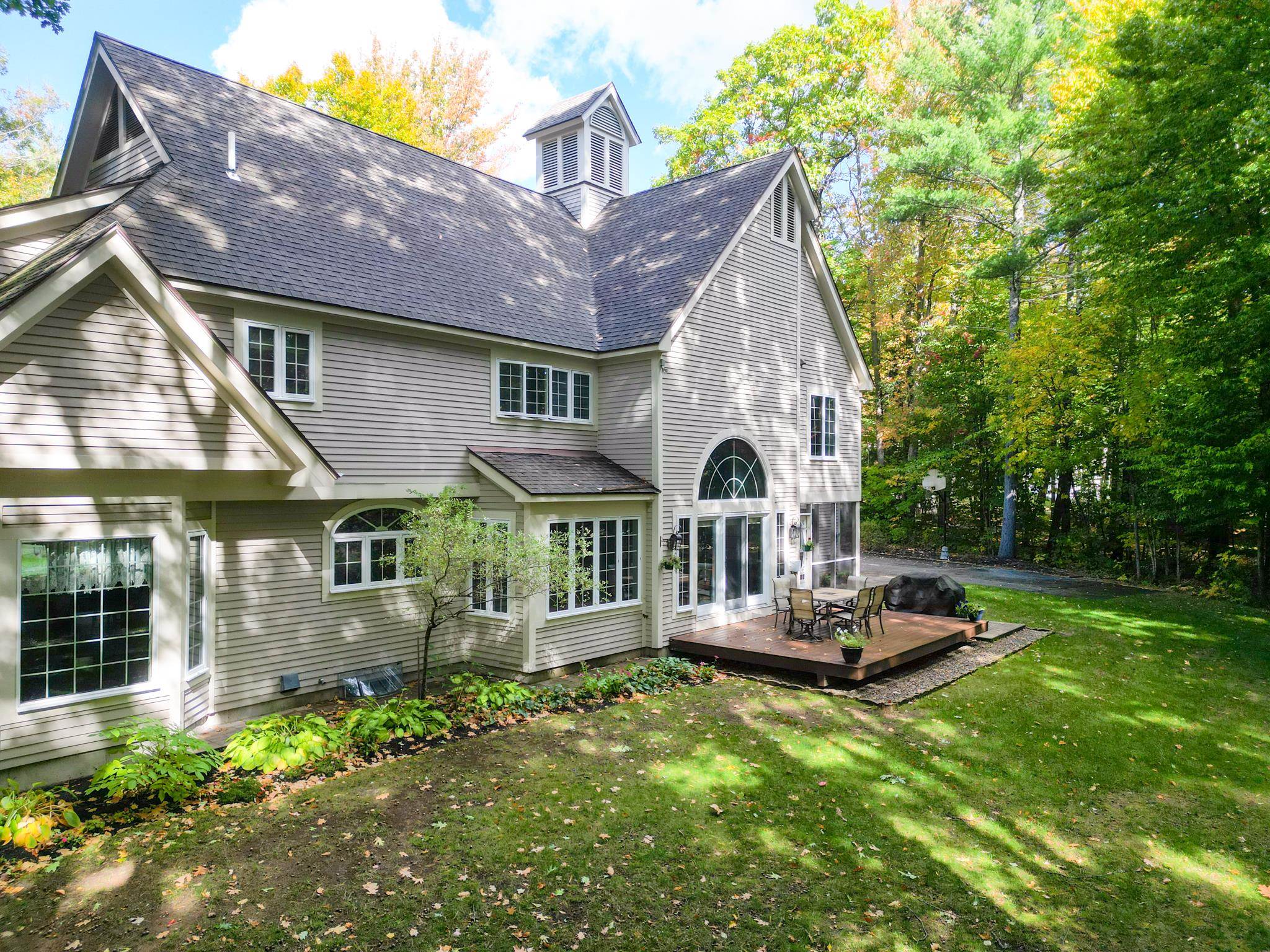 Rear exterior of a gray-sided home with an arched window and a deck among autumn trees.