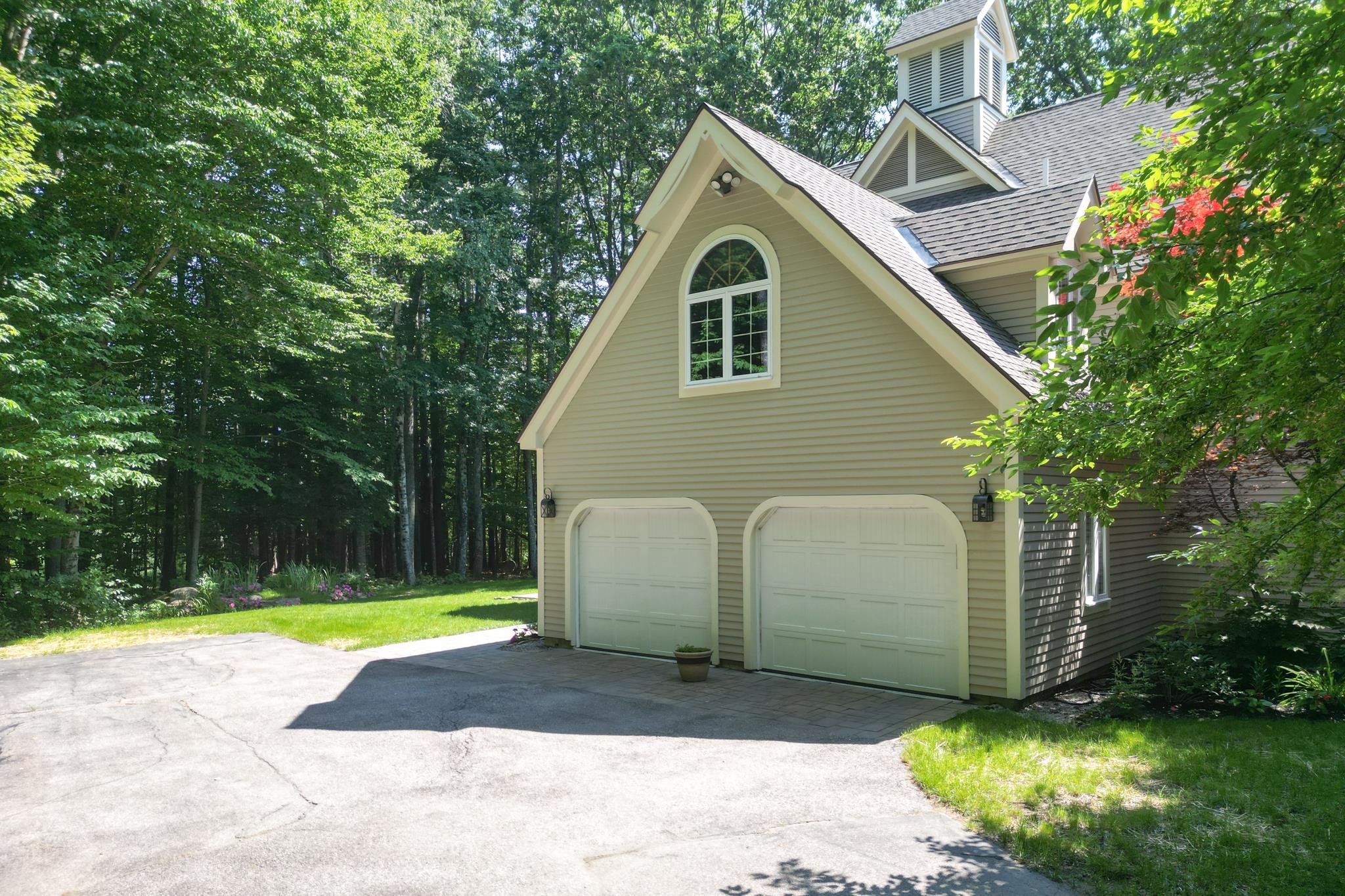 Detached beige garage with two white doors and a steep gabled roof, surrounded by leafy trees.