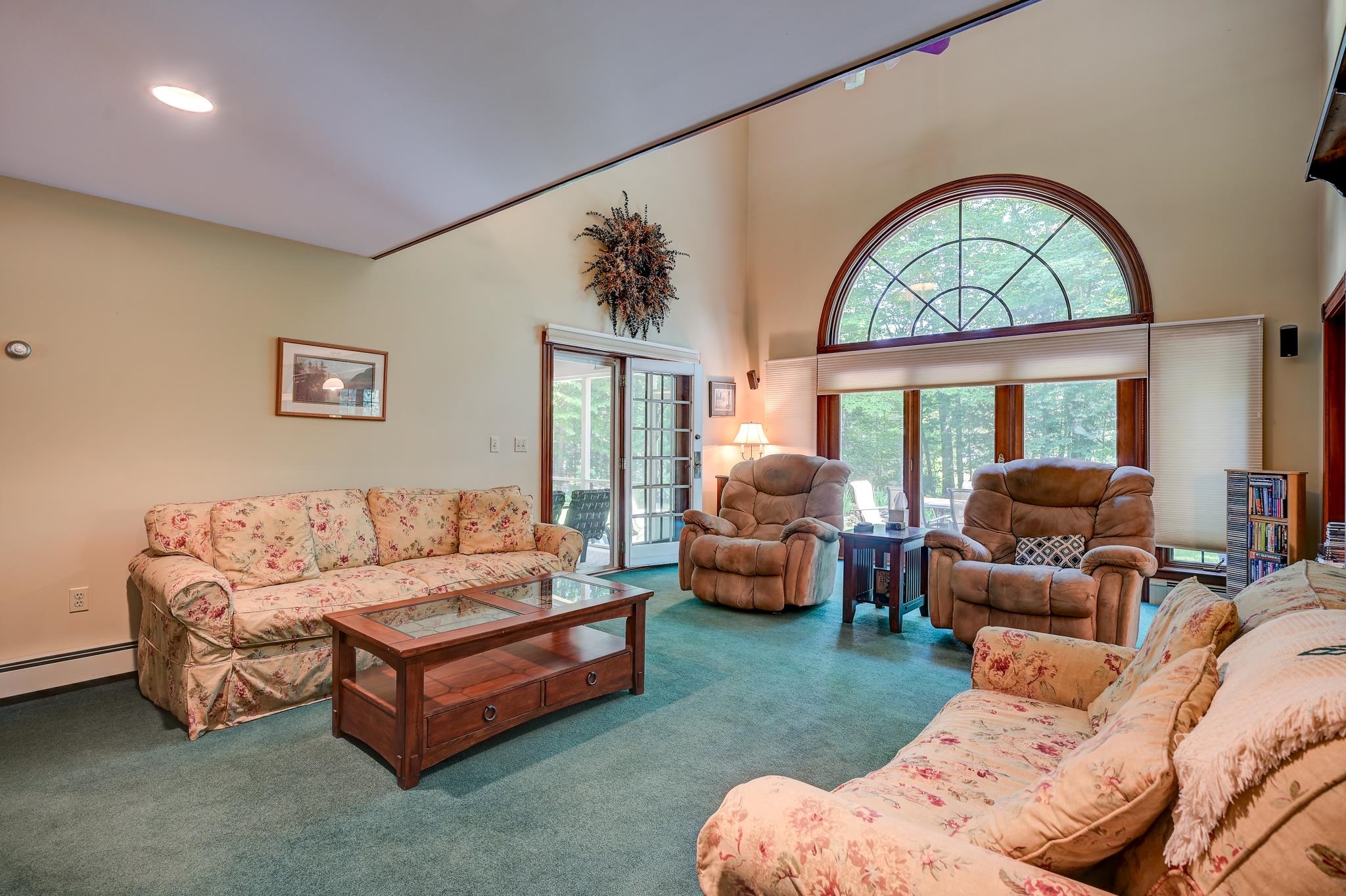 Furnished living room with floral sofas, recliners, and a large arched window overlooking trees.