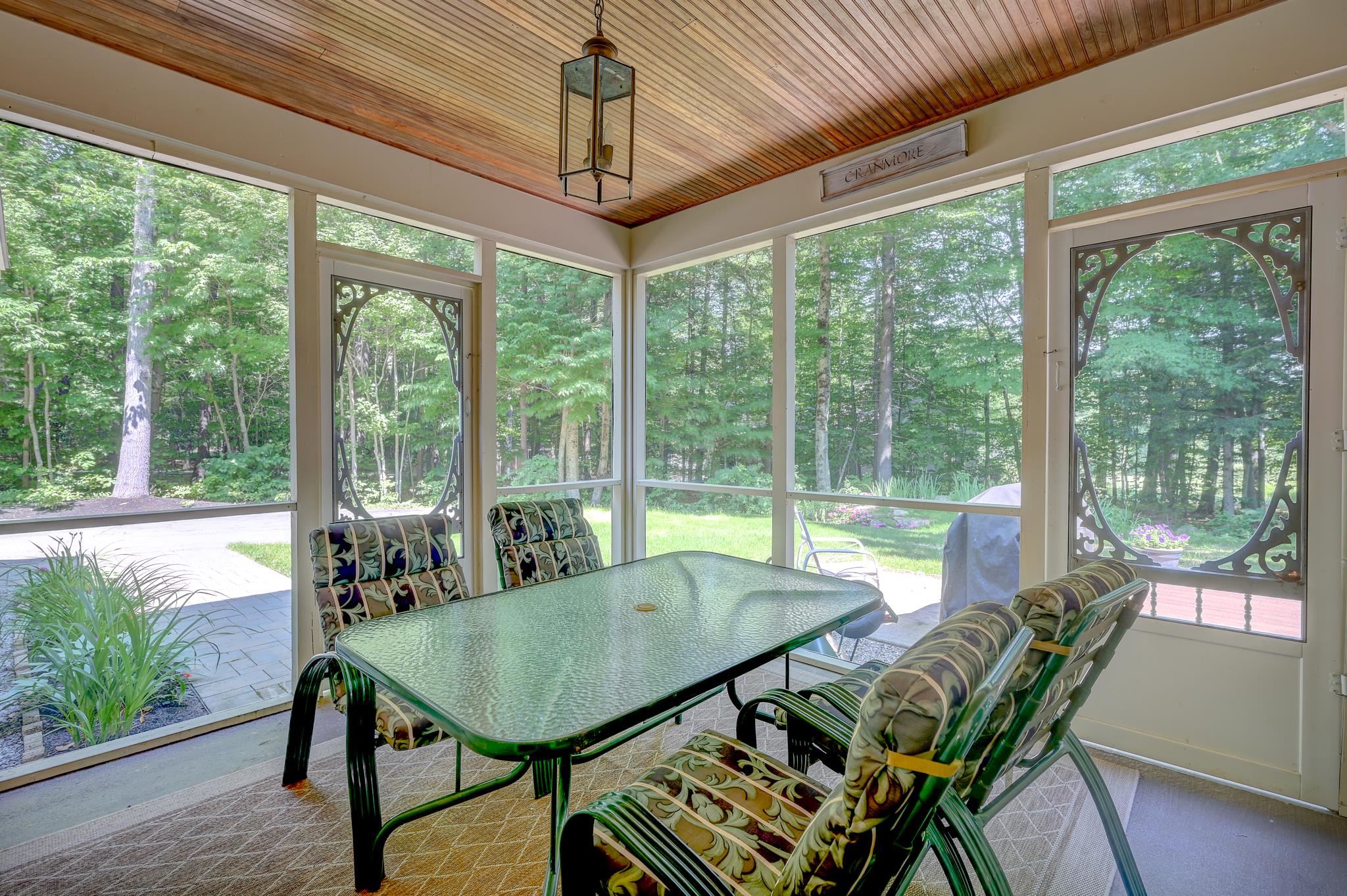 Enclosed sunroom with a glass dining table, patterned chairs, and wooded views through large windows.