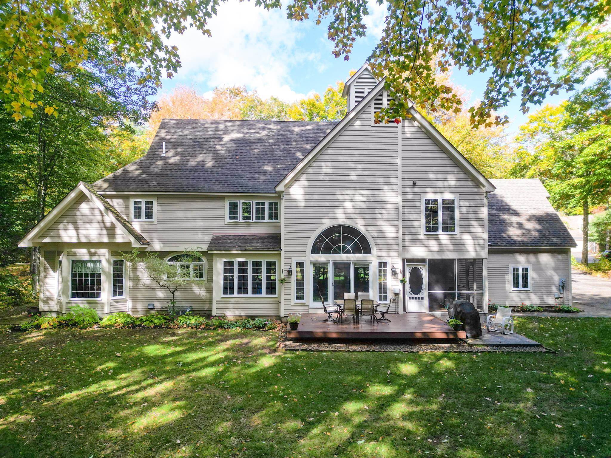 Rear exterior of a gray-sided home with a large arched window, deck, and leafy autumn trees.