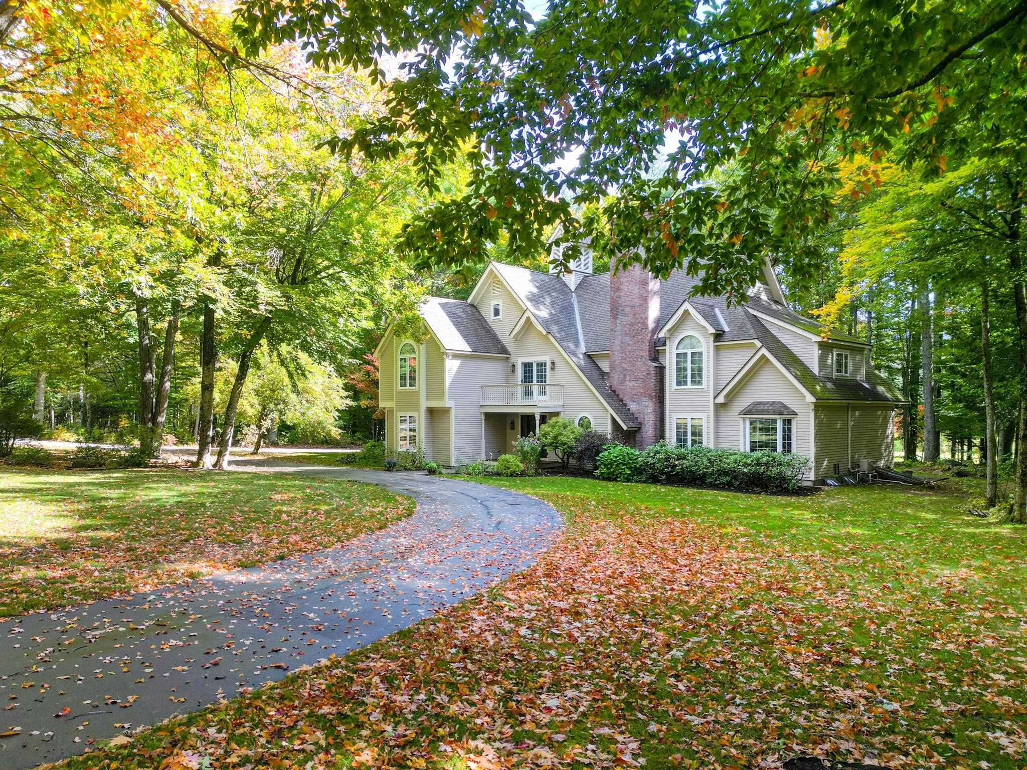 Large light-colored home with a brick chimney, framed by autumn trees and a curving driveway.