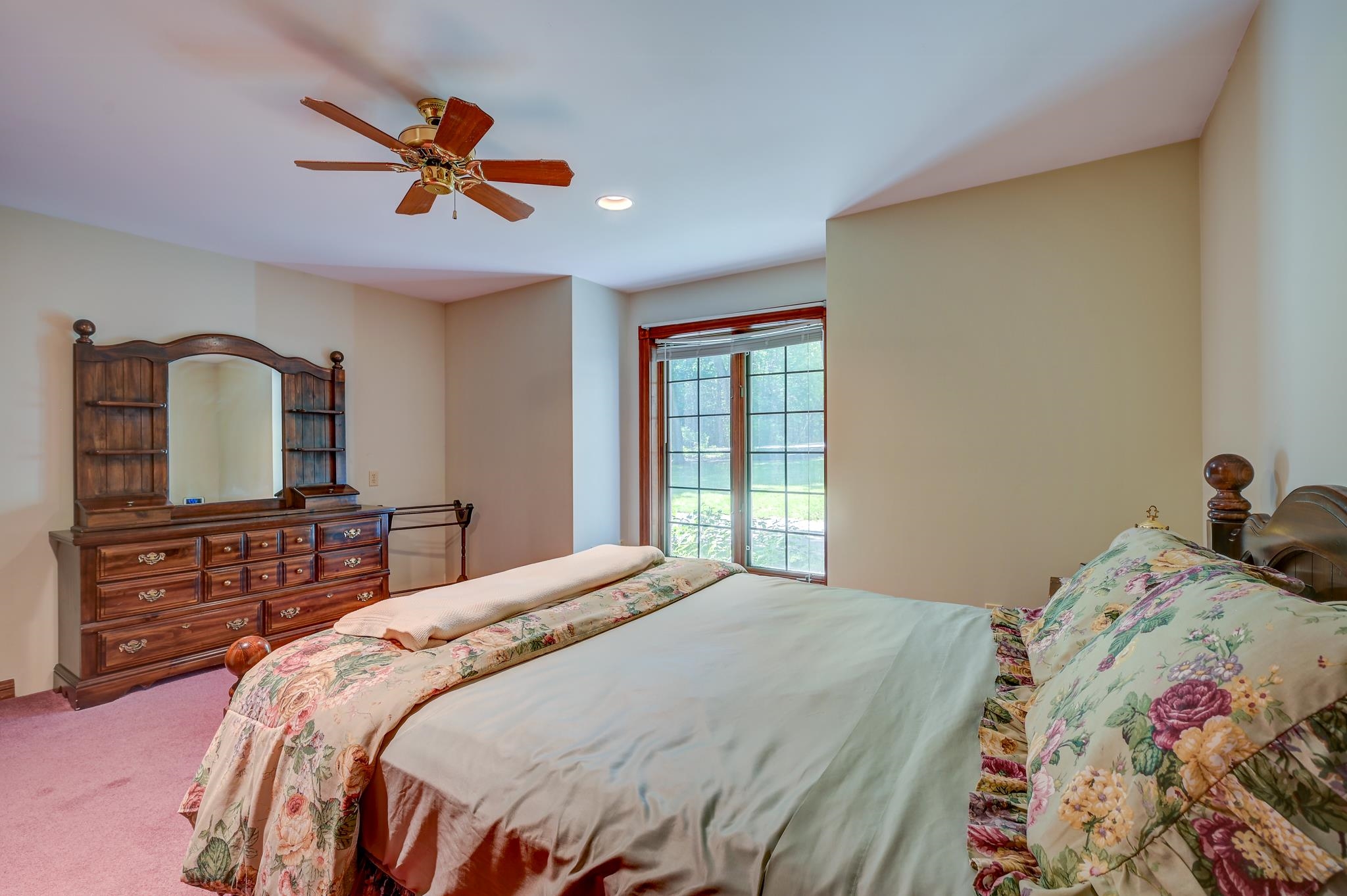 Furnished bedroom with a floral bedspread, dark wood dresser, and large window.