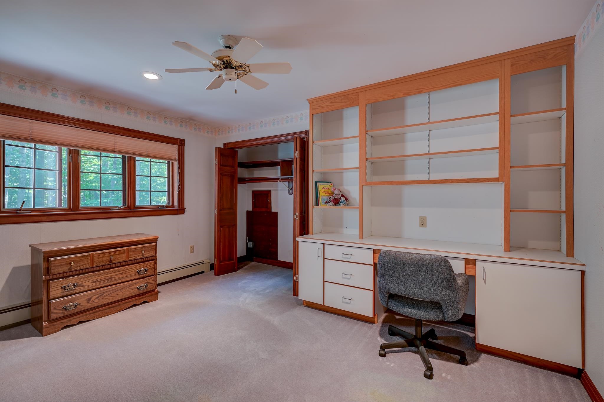 Bedroom with built-in desk and shelving, a wood dresser, and windows overlooking trees.