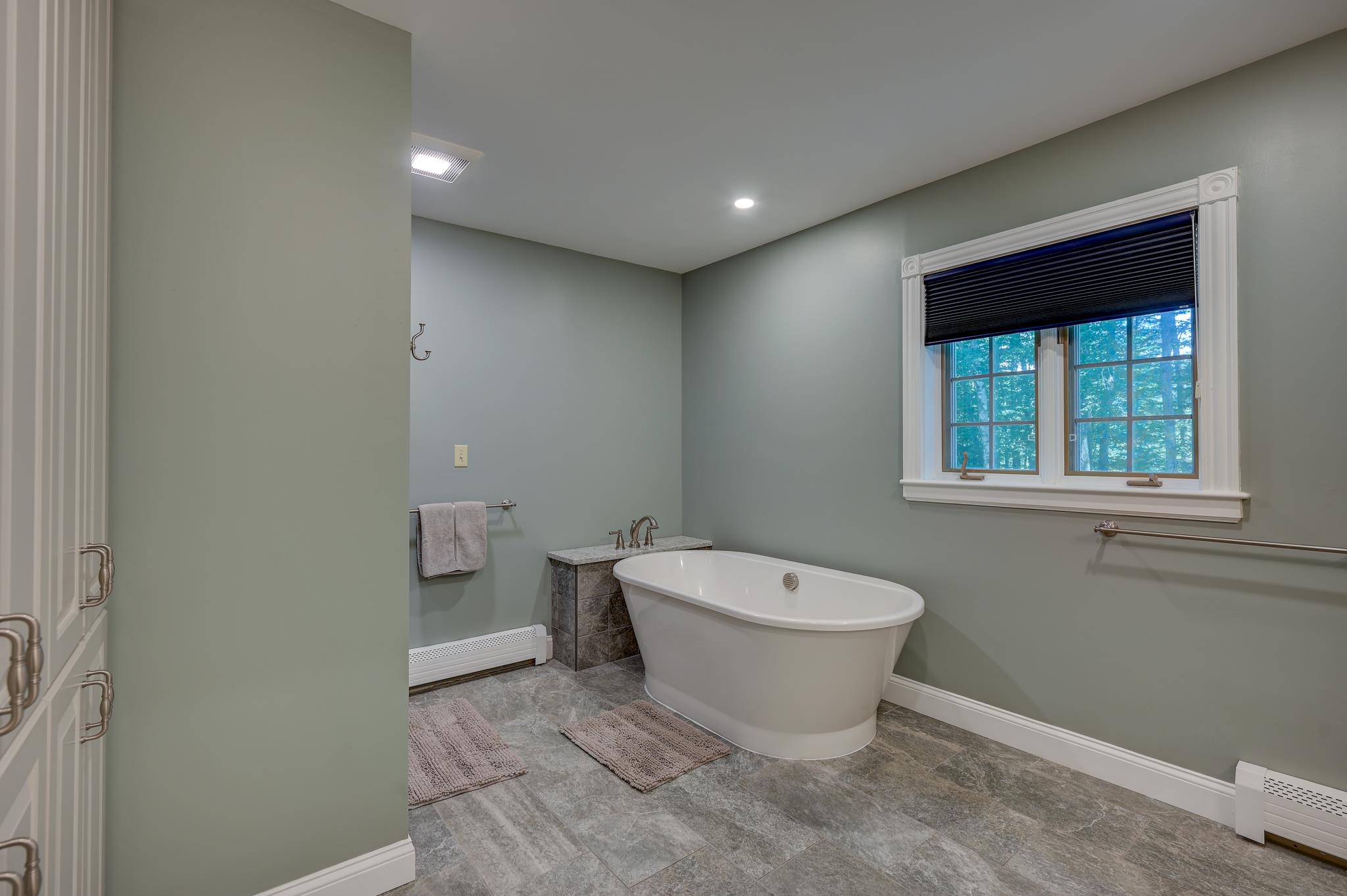 Bathroom with a freestanding soaking tub beneath a double window with dark blinds.