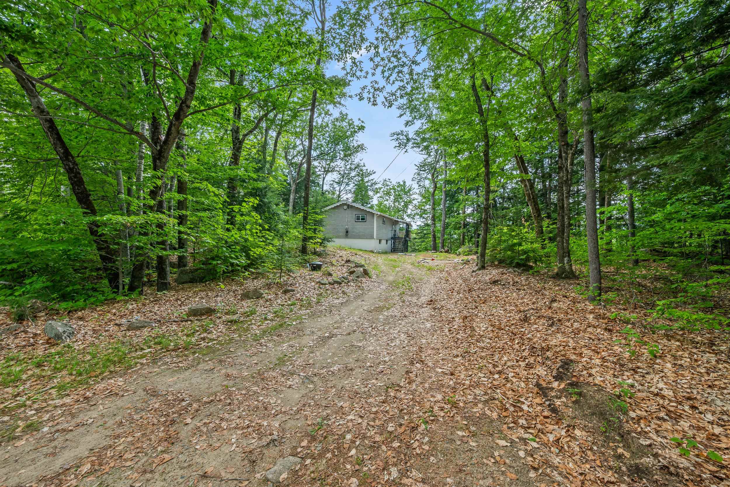 Gray raised home at the end of a wooded, leaf-covered driveway, with exterior stairs.