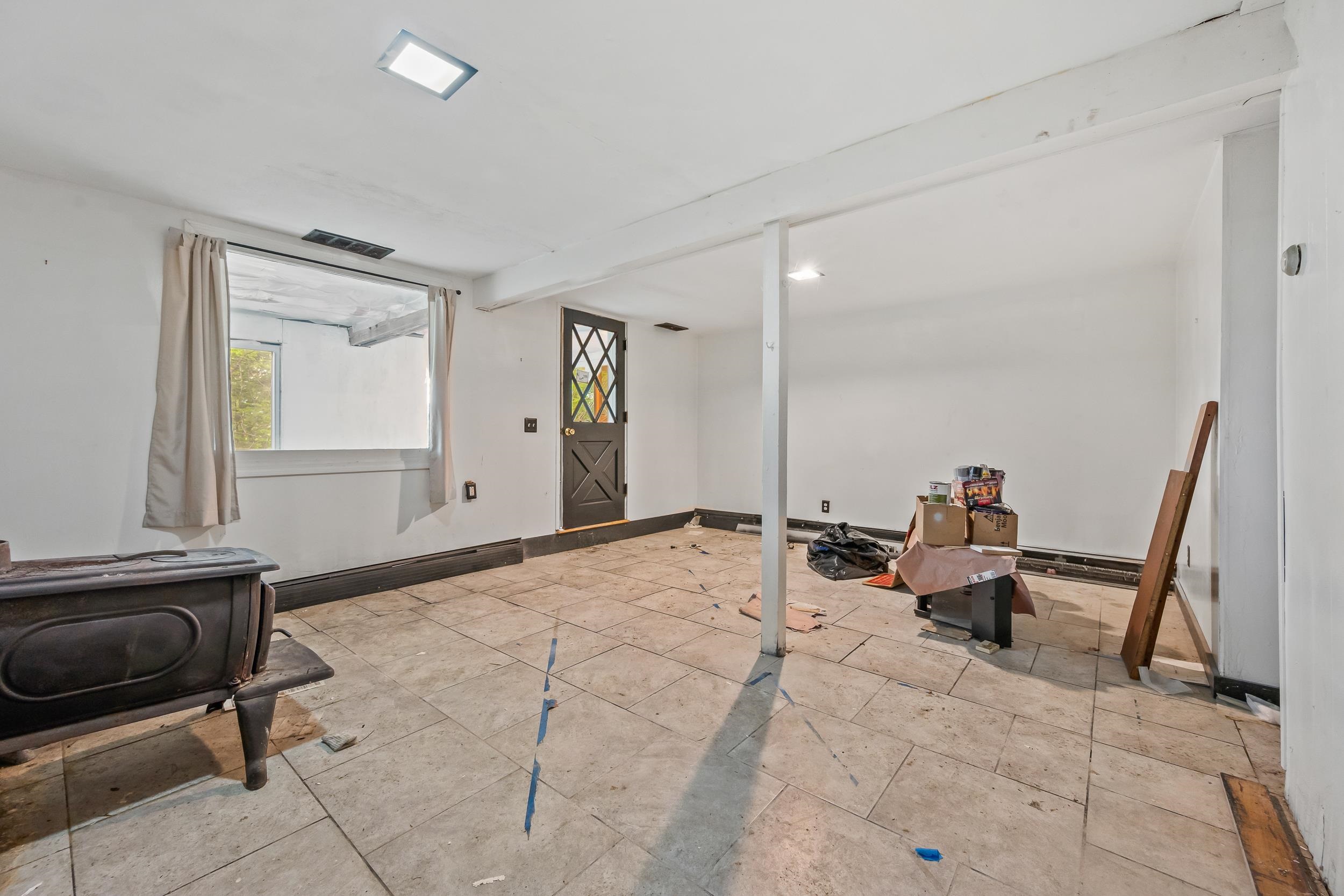 Unfinished tiled room with a wood stove, curtained window, and renovation materials.