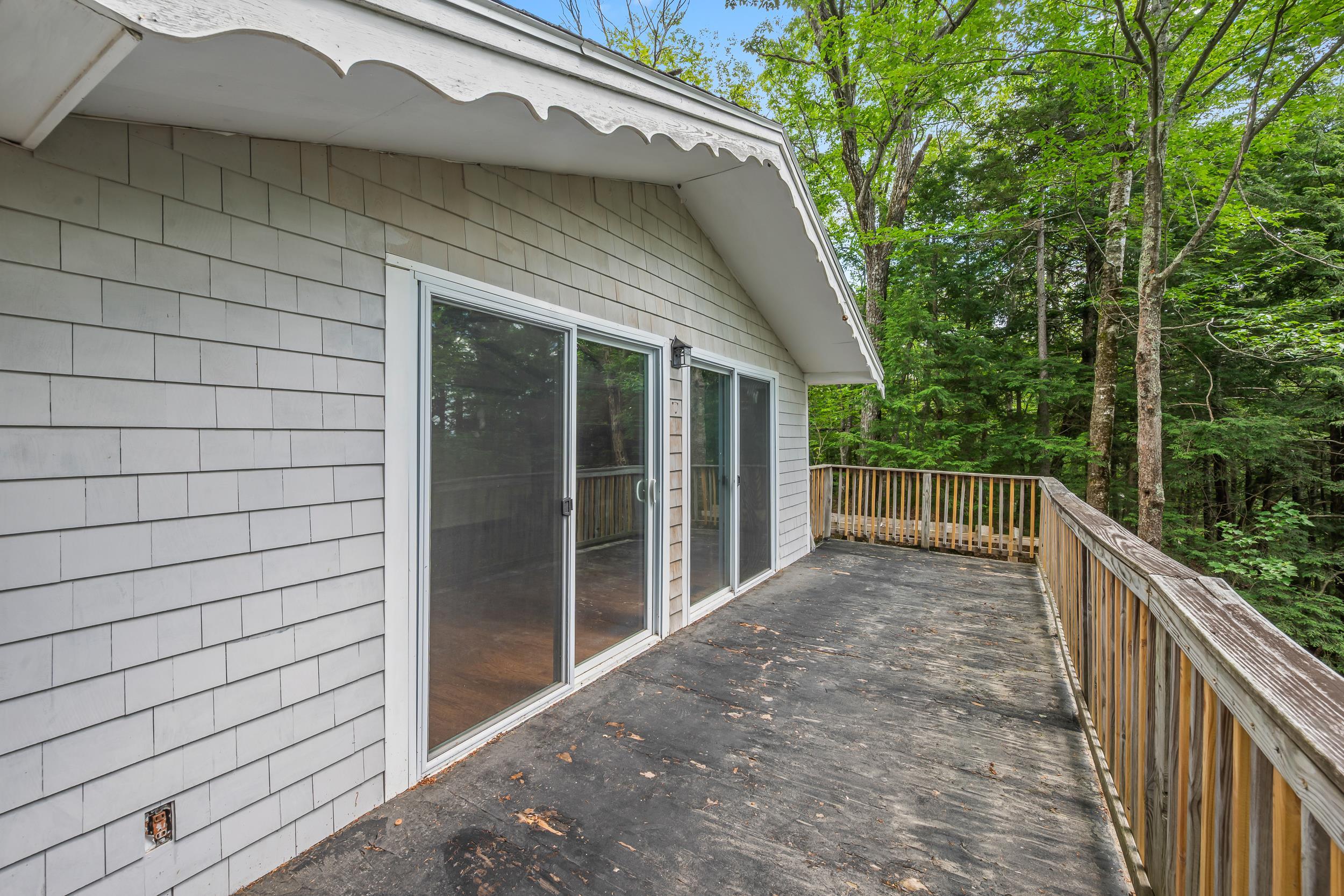 Wooden deck beside gray-shingled home, with sliding glass doors and a railing overlooking wooded surroundings.