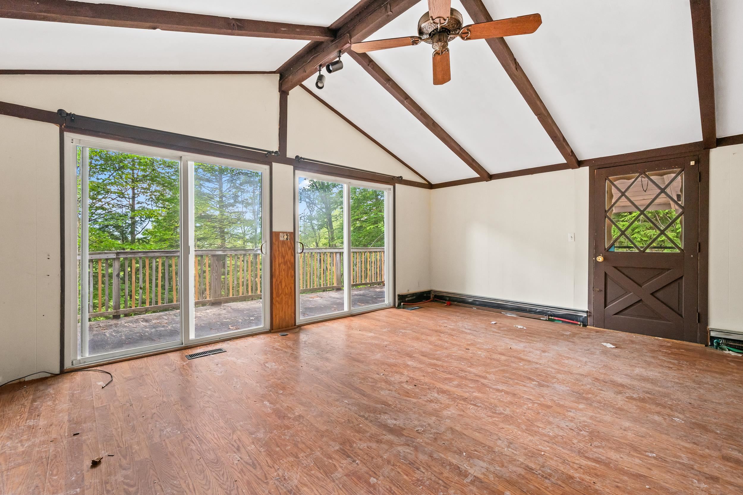 Empty vaulted room with exposed beams, sliding glass doors to a wooded deck, and a dark wood door.