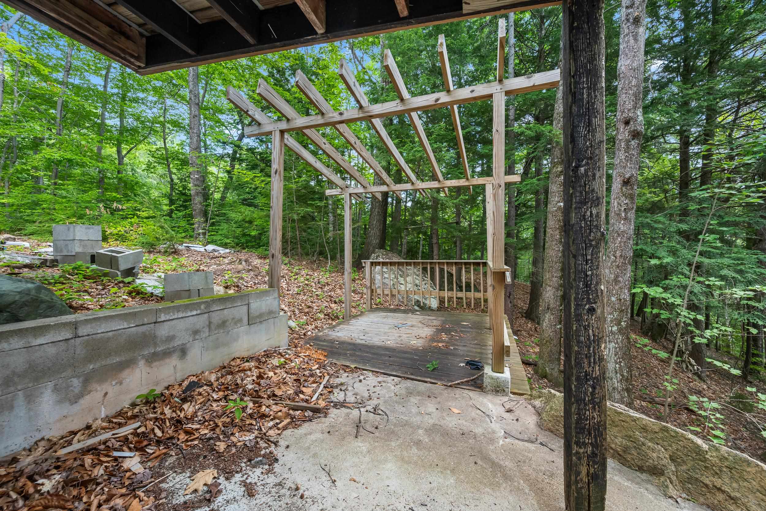 Concrete patio and unfinished wooden pergola and deck surrounded by trees.