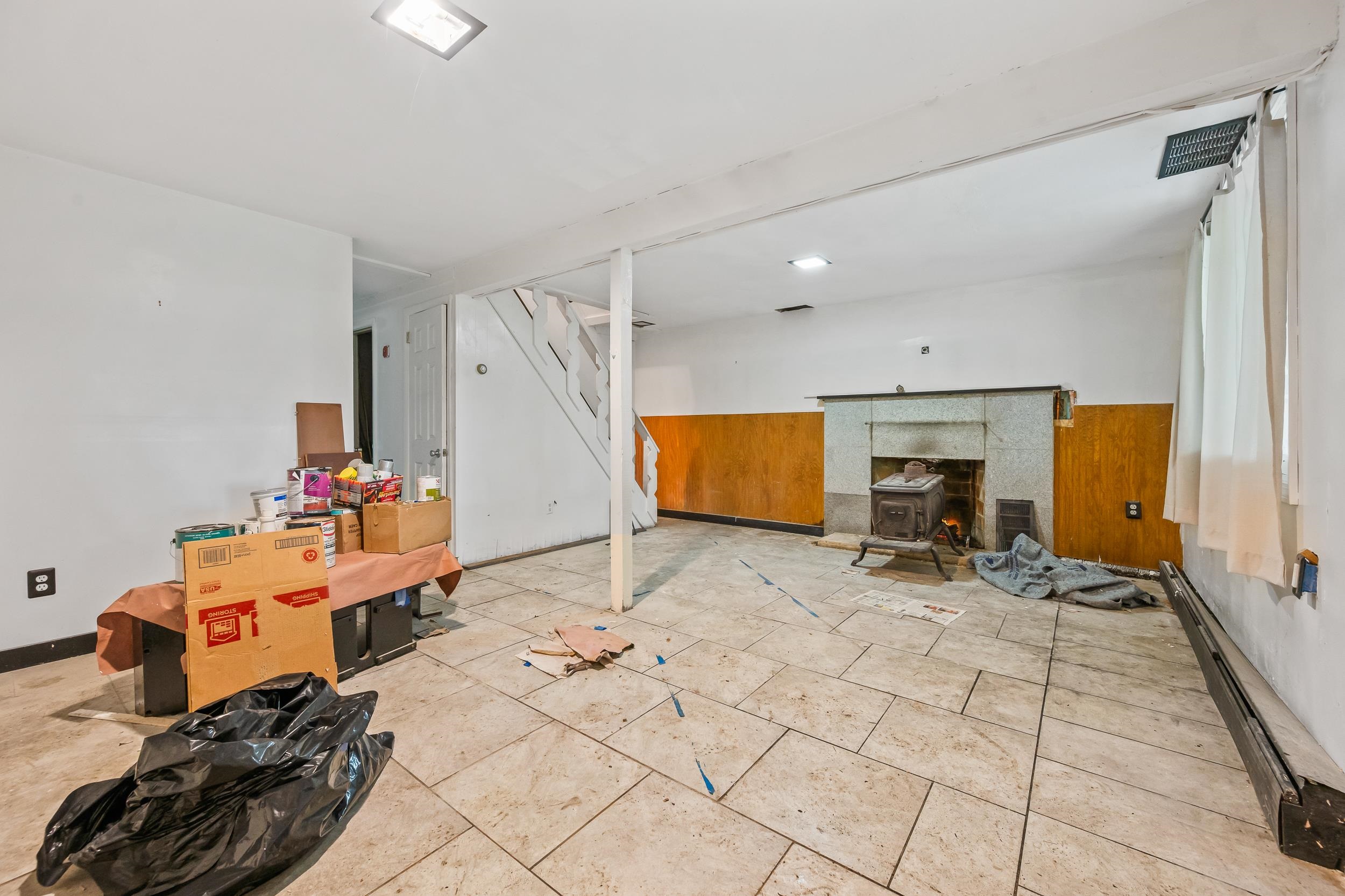 Lower-level room with tiled floor, staircase, and wood stove amid renovation supplies and debris.