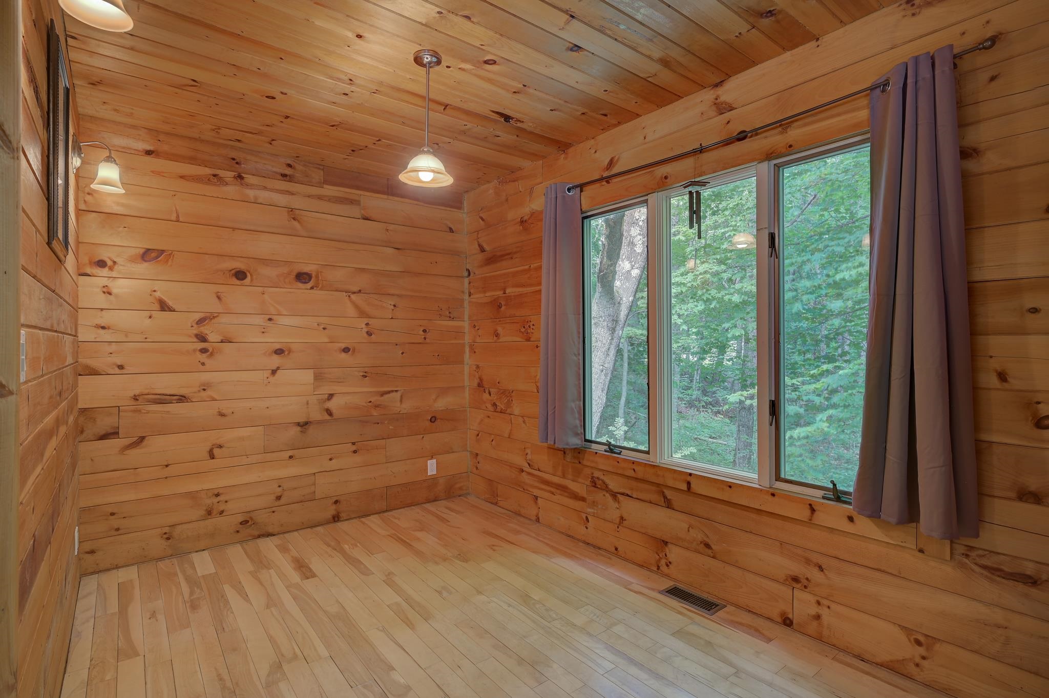 Empty wood-paneled room with light plank flooring and a large window overlooking trees.