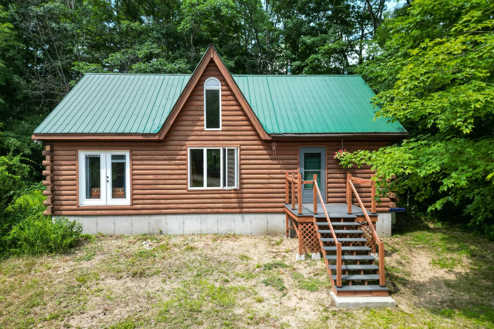 Log cabin with a green metal roof, raised porch, and stairs among leafy trees.