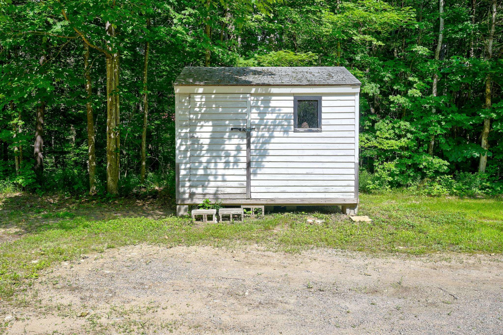 White-sided shed with a dark roof in a wooded clearing beside a gravel area.