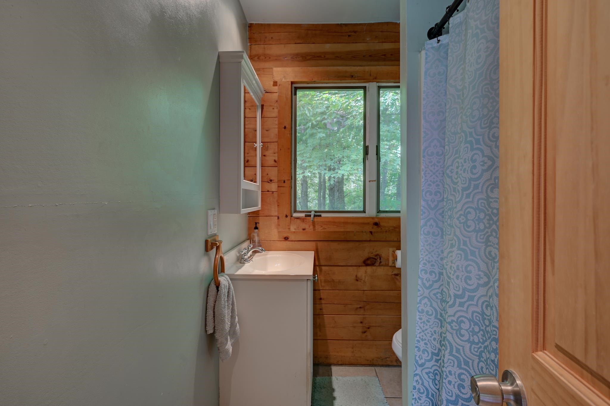 Bathroom with a white vanity, wood-plank walls, and a window overlooking trees.