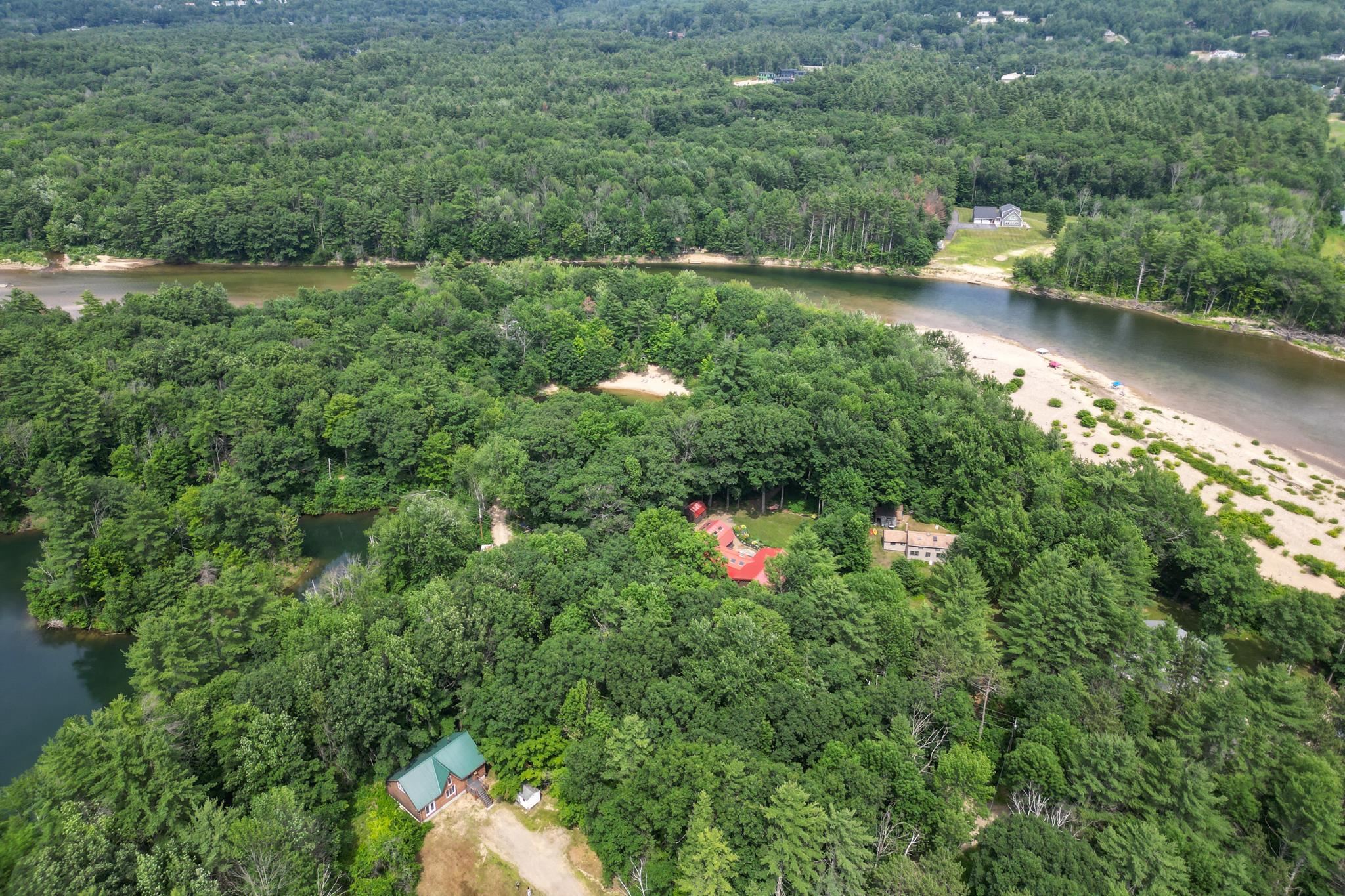 Aerial view of a green-roofed cabin amid forest, near a river and sandy beach.