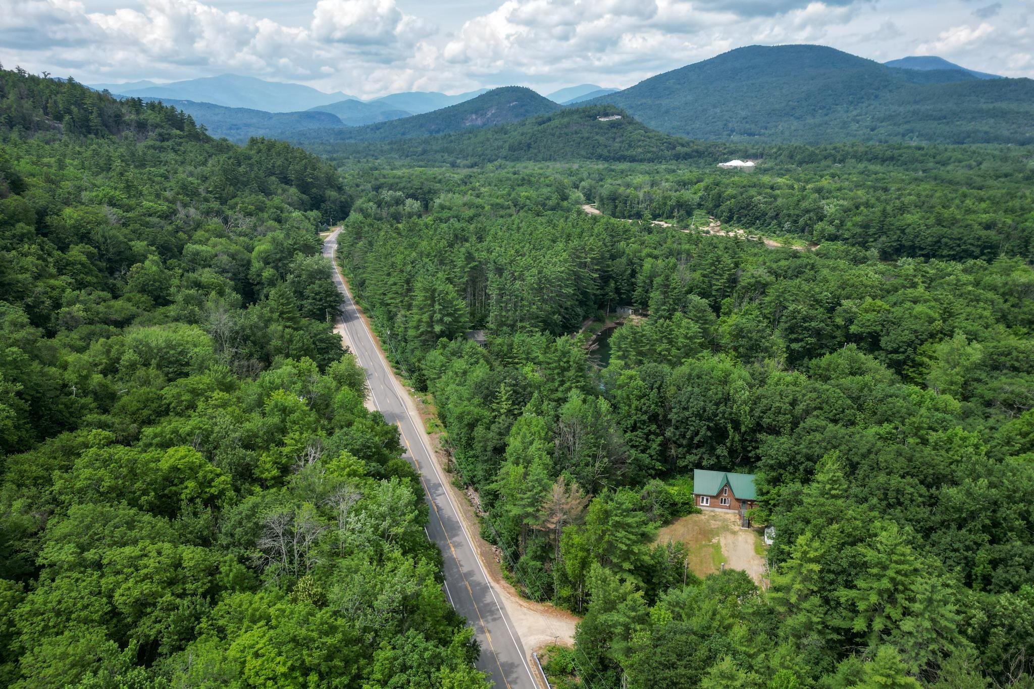 Aerial view of a green-roofed house amid dense forest, with a road and mountains beyond.