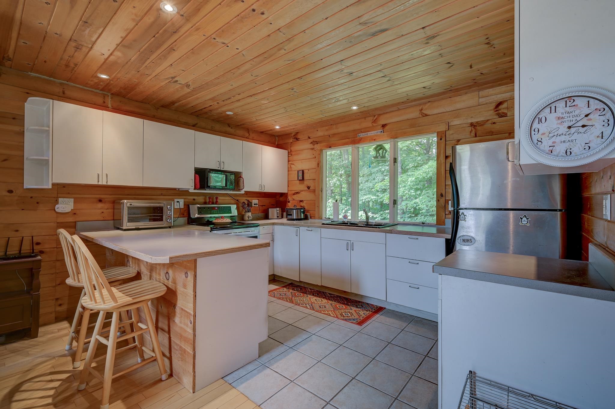 Kitchen with pine walls, white cabinets, a peninsula with stools, and a wide window overlooking trees.