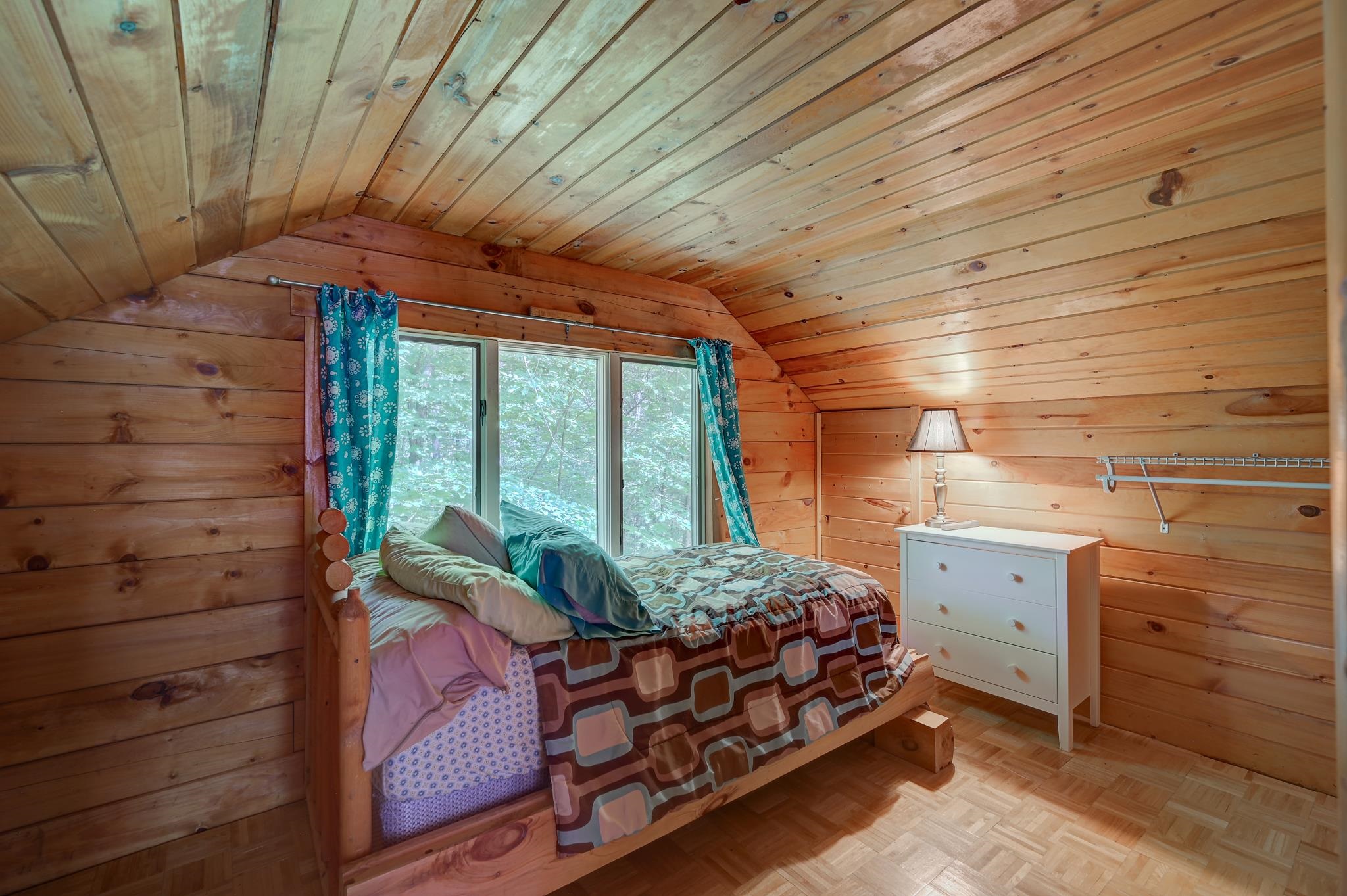 Cabin bedroom with a log bed beneath a sloped wood ceiling and a window facing trees.