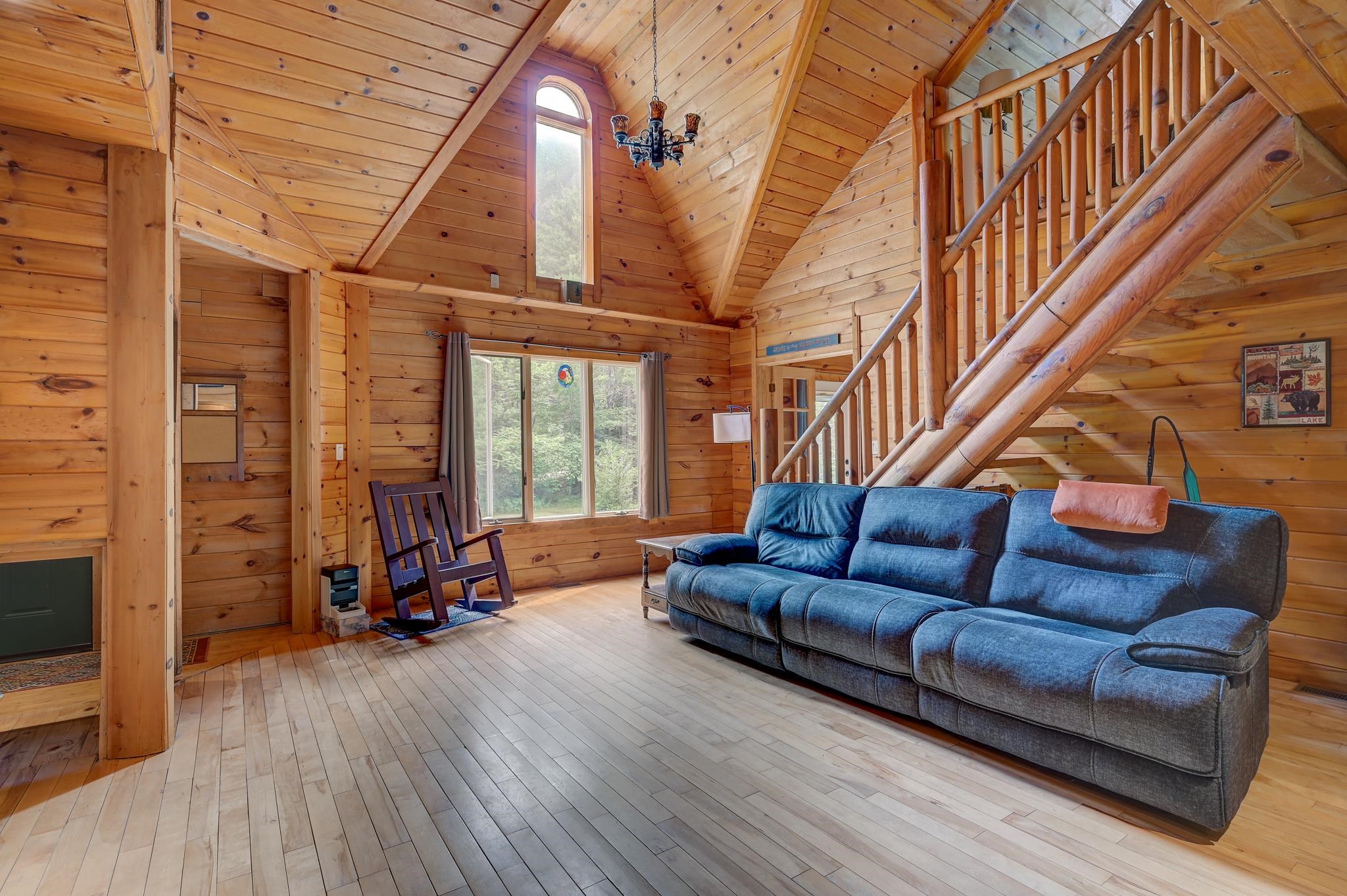 Cabin living room with vaulted wood ceiling, blue sofa, rocking chair, and log-railed staircase.