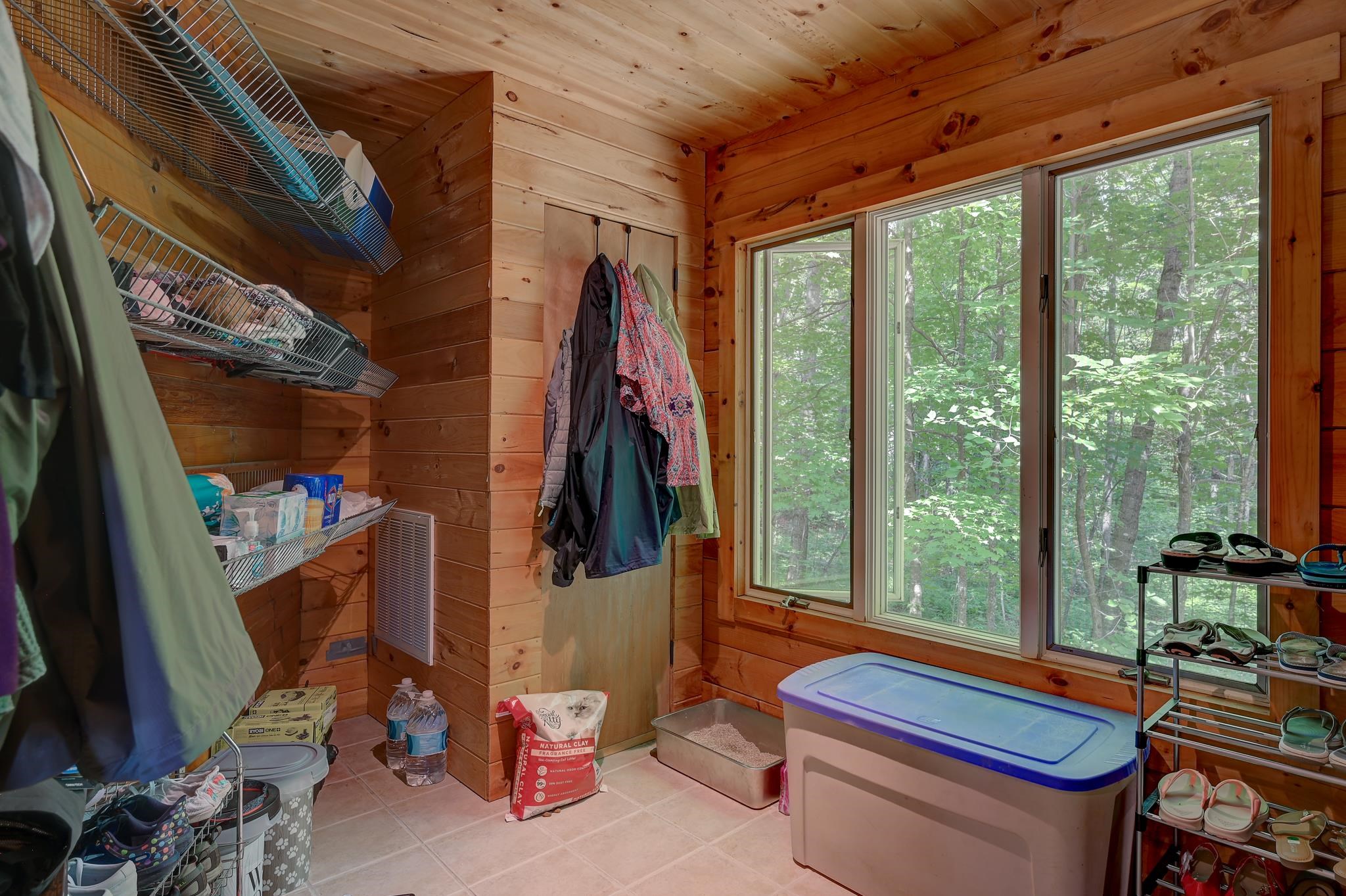 Wood-paneled mudroom with storage shelves, hanging coats, and a large window overlooking woods.