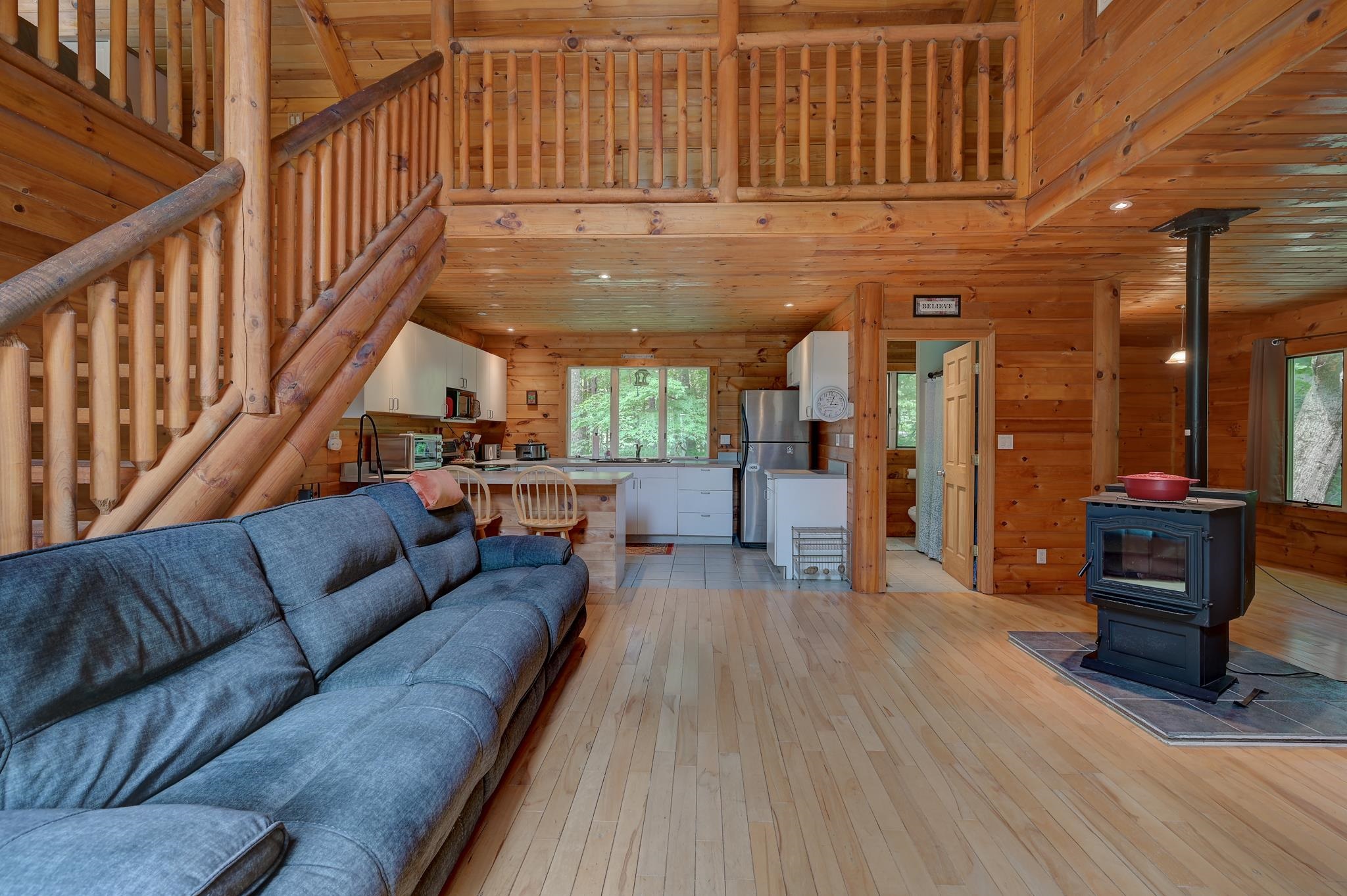 Cabin living room with a loft staircase, blue sectional, open kitchen, and black wood stove.