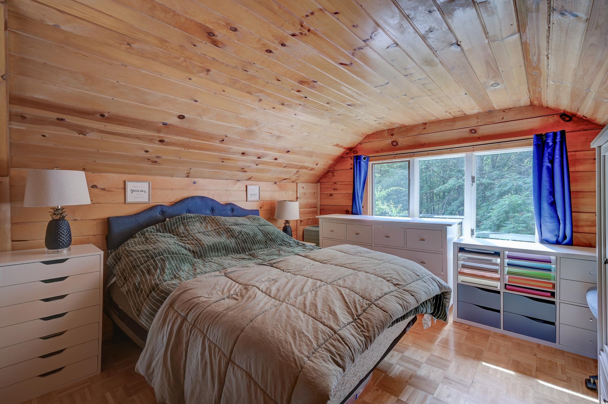 Bedroom with a sloped pine ceiling, blue curtains, and a wide window overlooking trees.