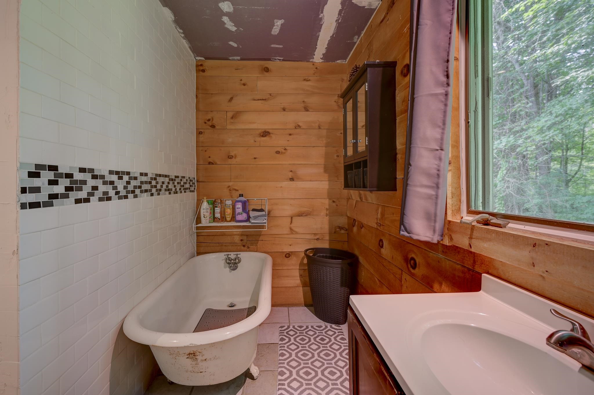 Bathroom with a freestanding tub, wood-plank walls, tiled accent band, and a large window overlooking trees.