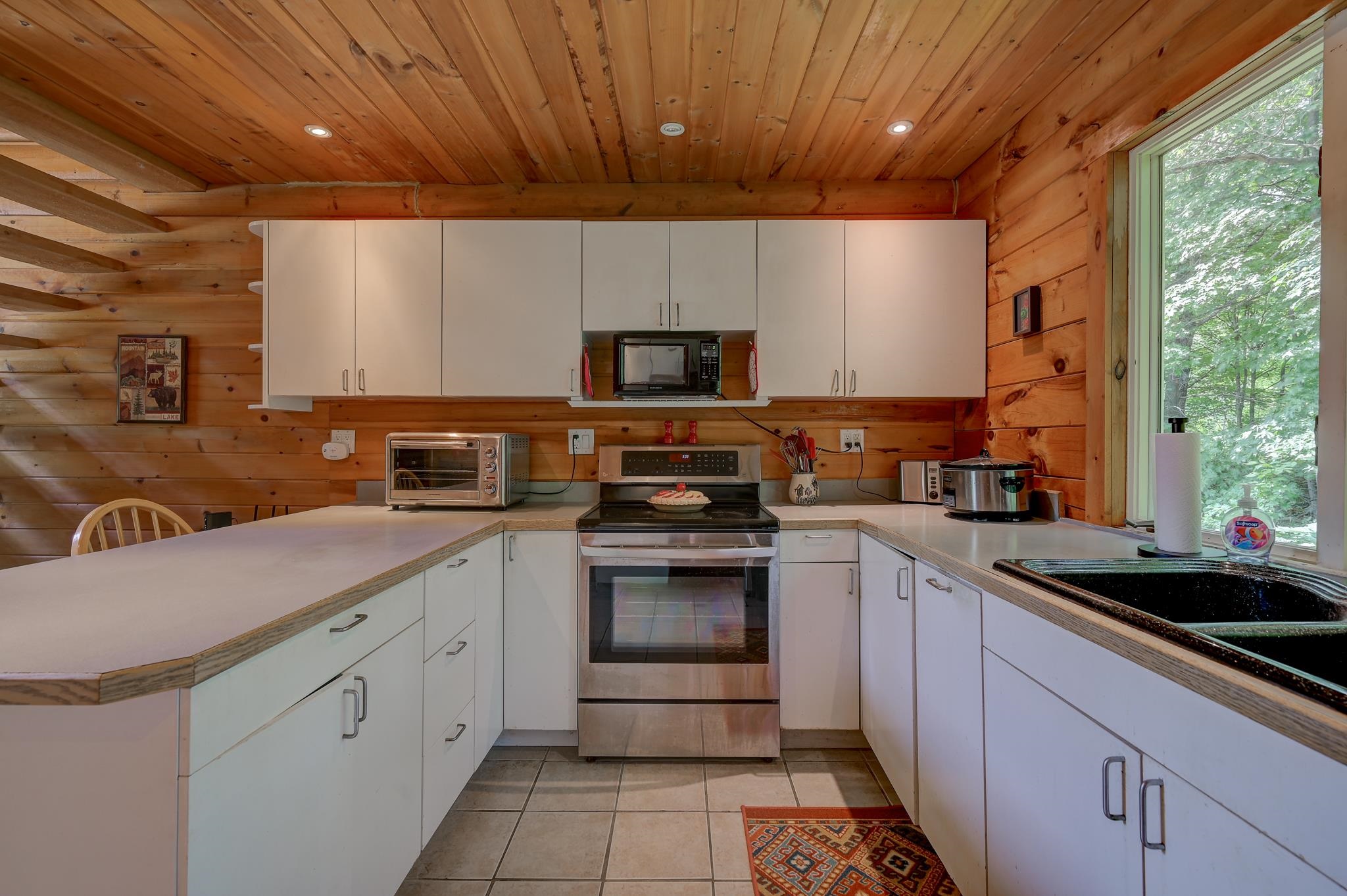 Kitchen with white cabinets, stainless steel range, warm wood walls, and a large window overlooking trees.