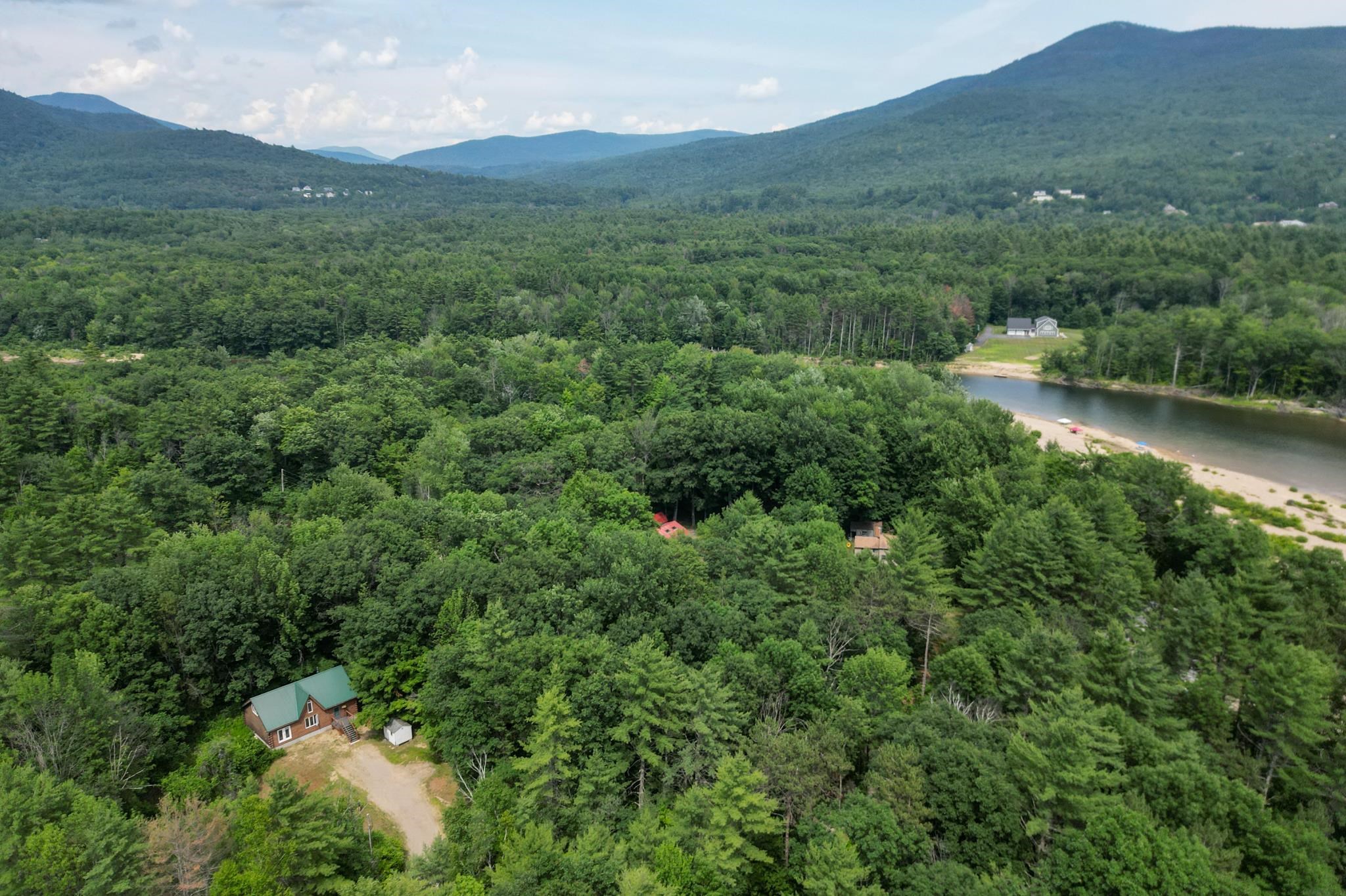 Aerial view of a cabin amid forest, with a sandy beach, lake, and mountains beyond.