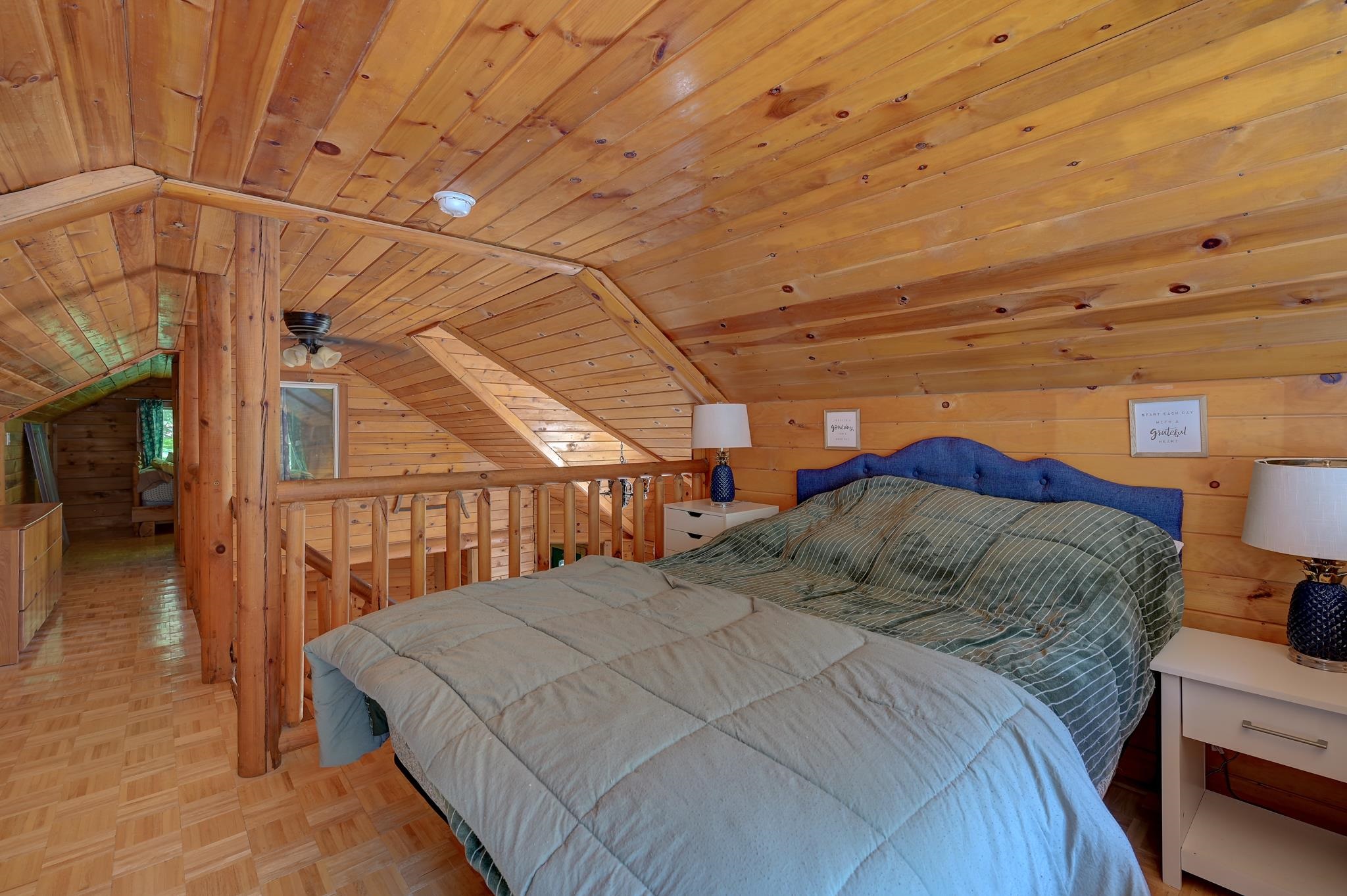Loft bedroom with a large bed beneath a knotty-pine ceiling and beside a wooden railing.