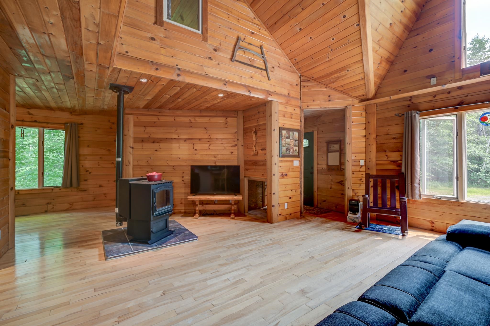Vaulted pine living room with a wood stove, blue sofa, and windows overlooking trees.