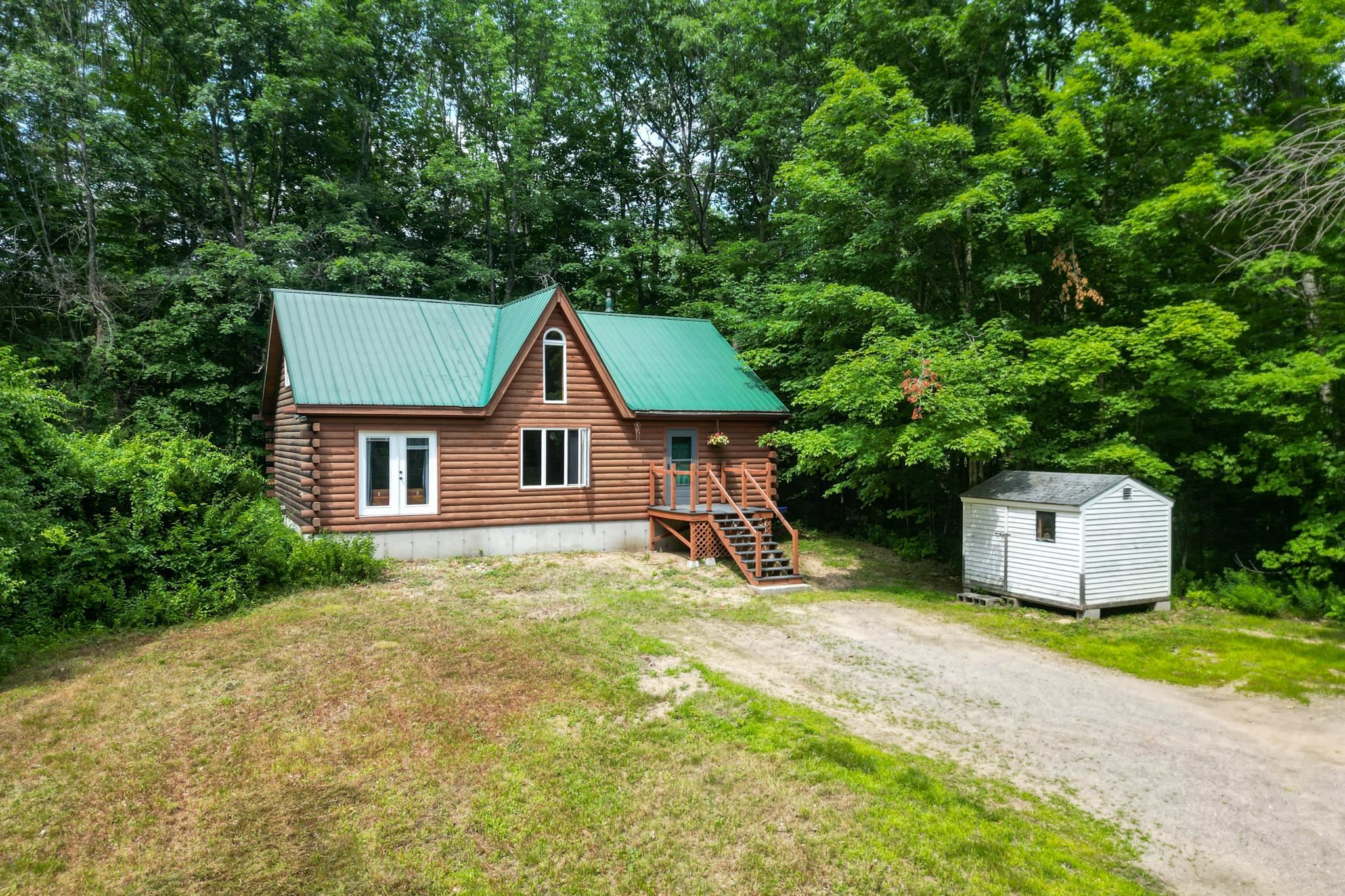 Log cabin with a green metal roof and front porch, beside a white shed in a wooded setting.
