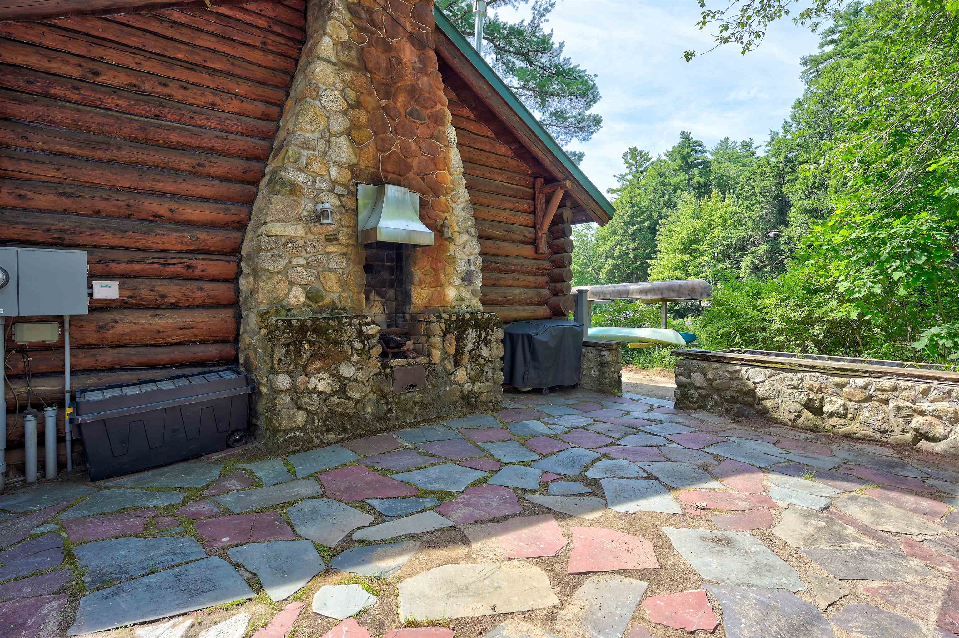 Log home patio with a large stone fireplace, flagstone paving, and wooded surroundings.