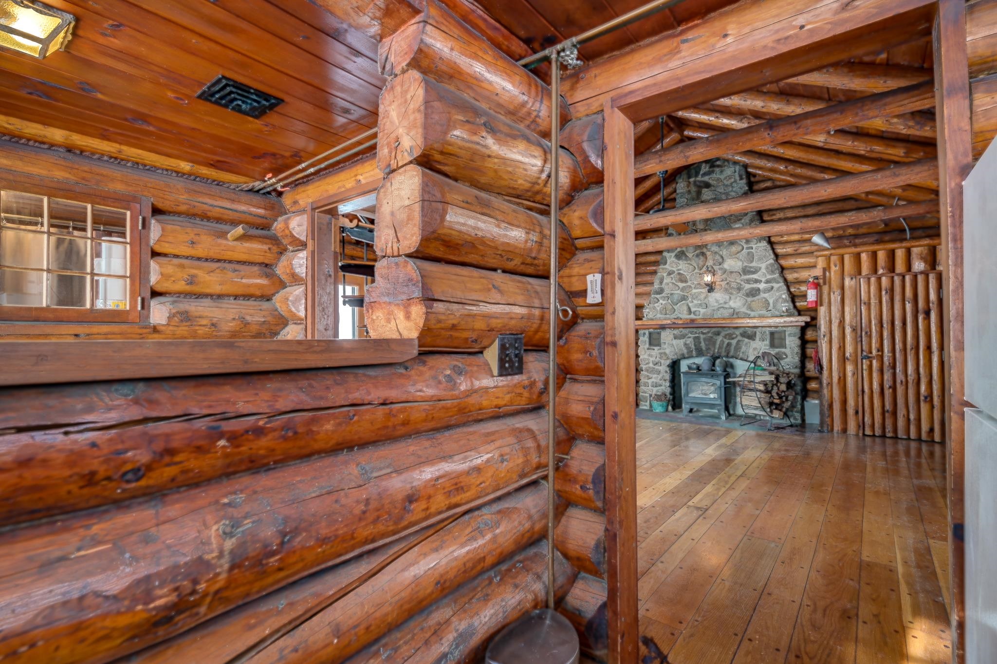 Log cabin interior with exposed timber beams, plank floors, and a stone fireplace with a wood stove.