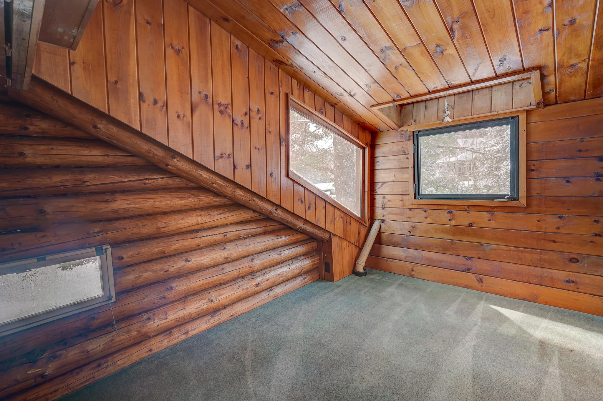 Empty cabin room with a sloped wood ceiling, exposed log wall, gray carpet, and snowy views through the windows.