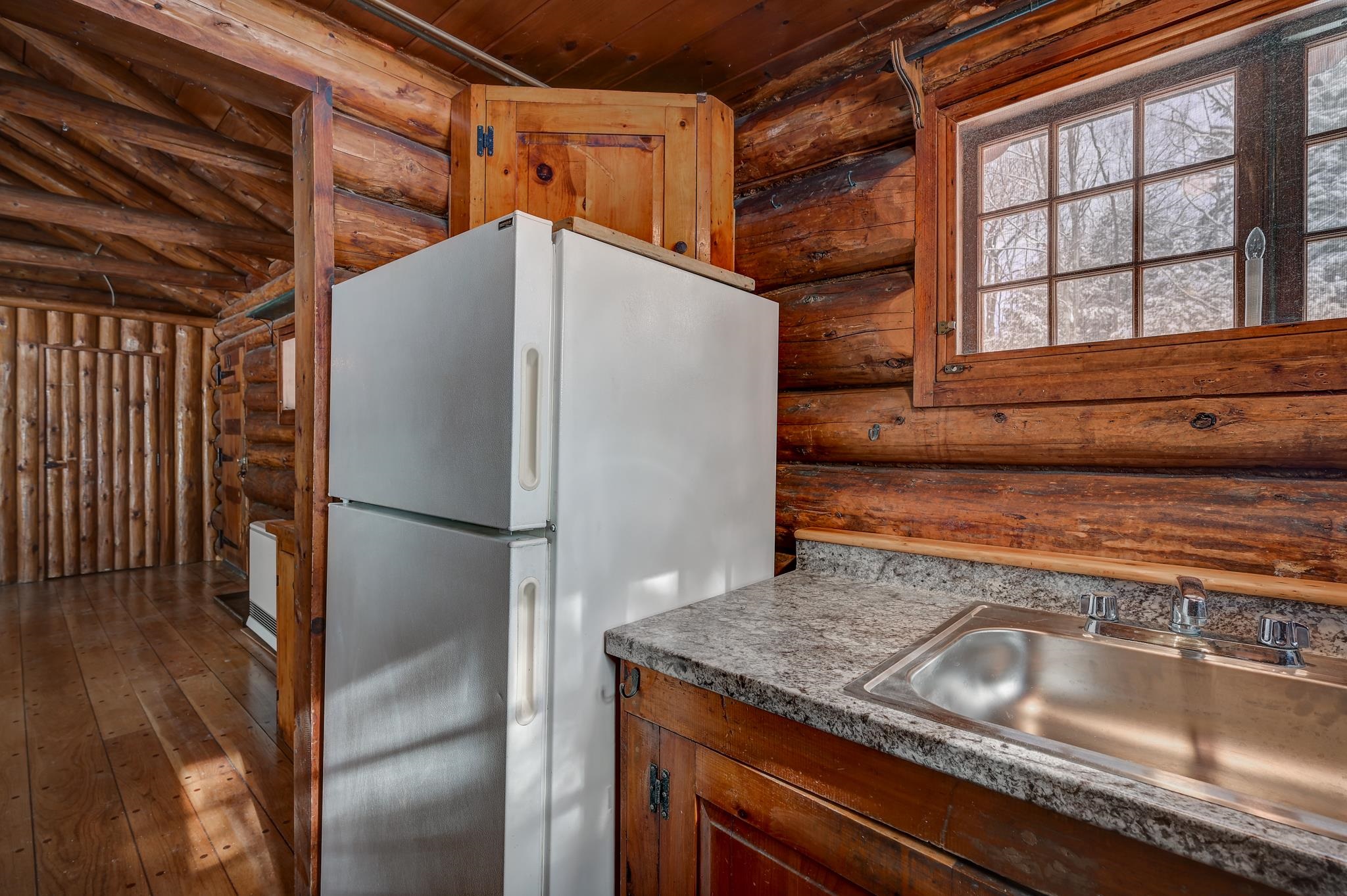 Rustic log cabin kitchen with a white refrigerator, granite counter, stainless-steel sink, and multi-pane window.