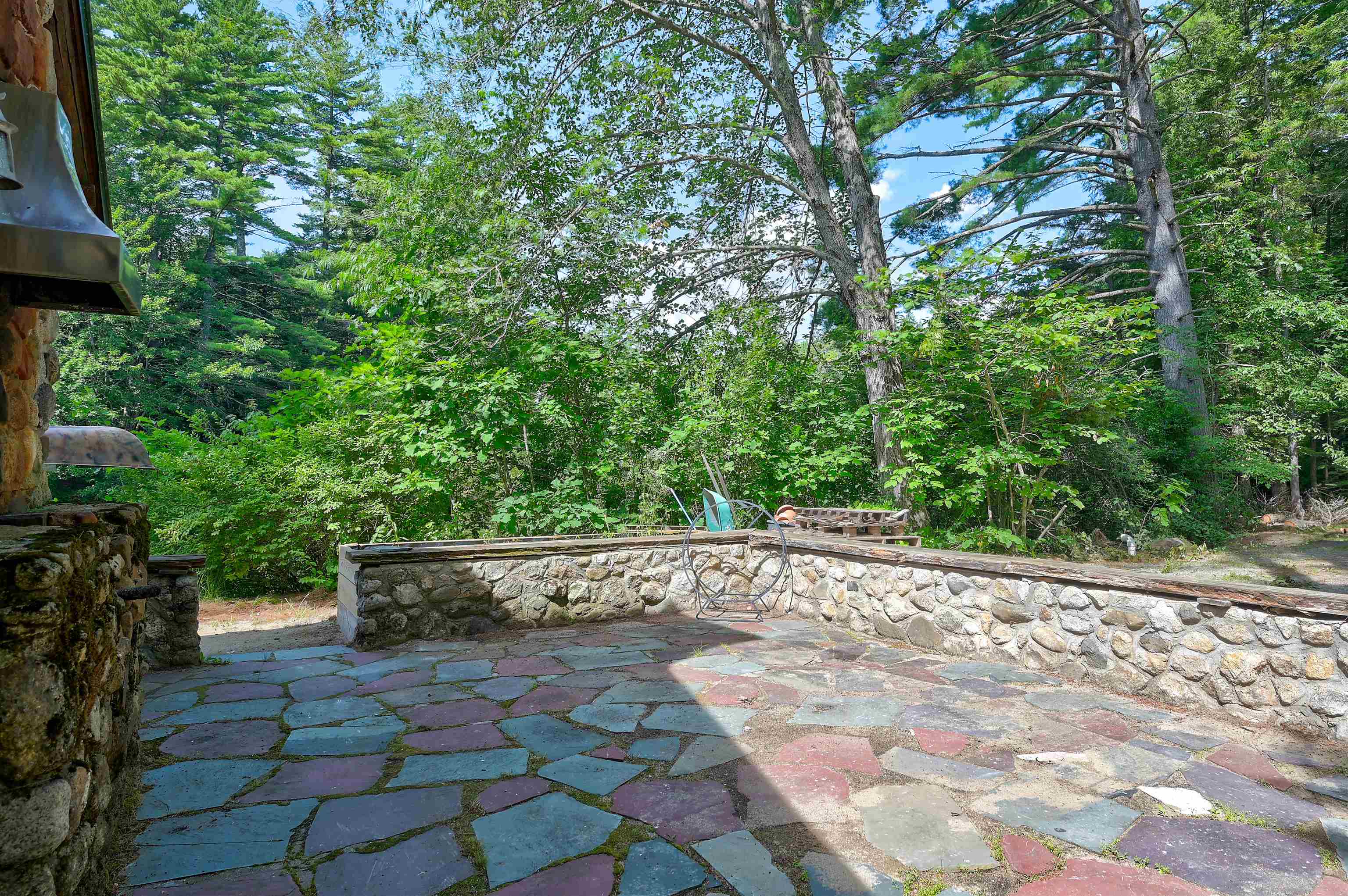 Flagstone patio with a low stone wall and metal hanging chair, surrounded by trees.