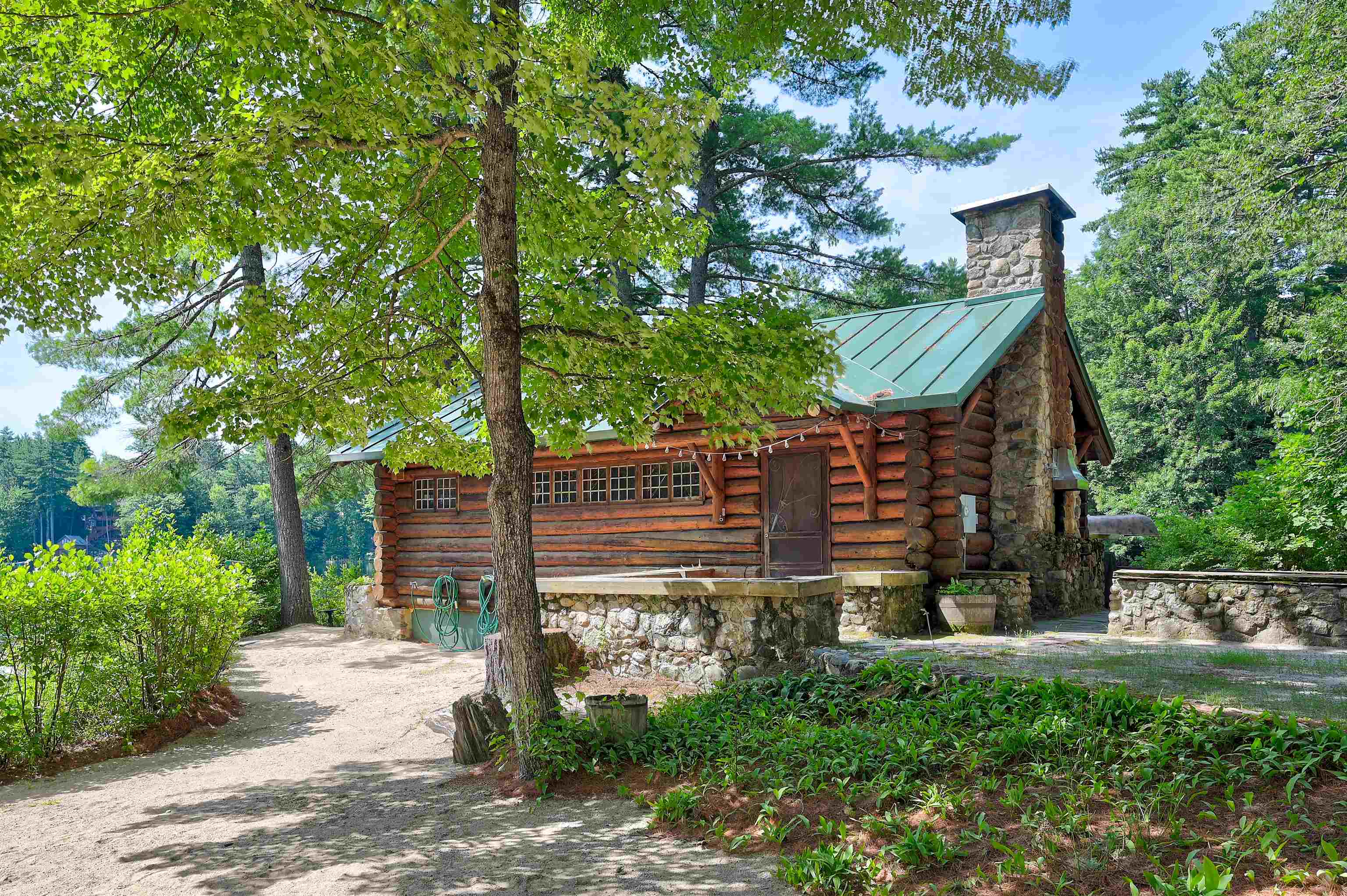 Log cabin with a green metal roof and stone chimney among mature trees.