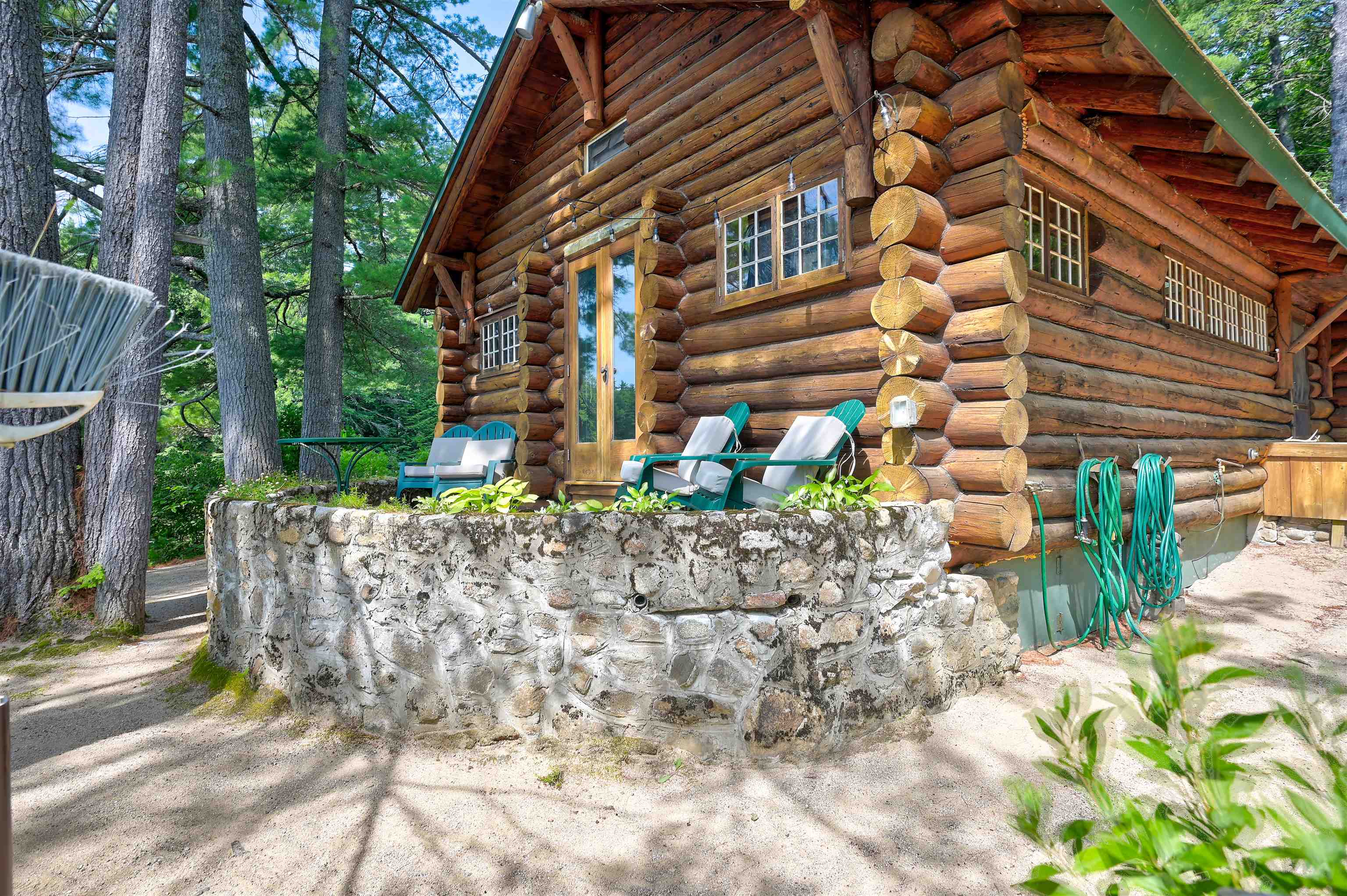 Log cabin exterior with a stone terrace, outdoor chairs, and a wooded setting.