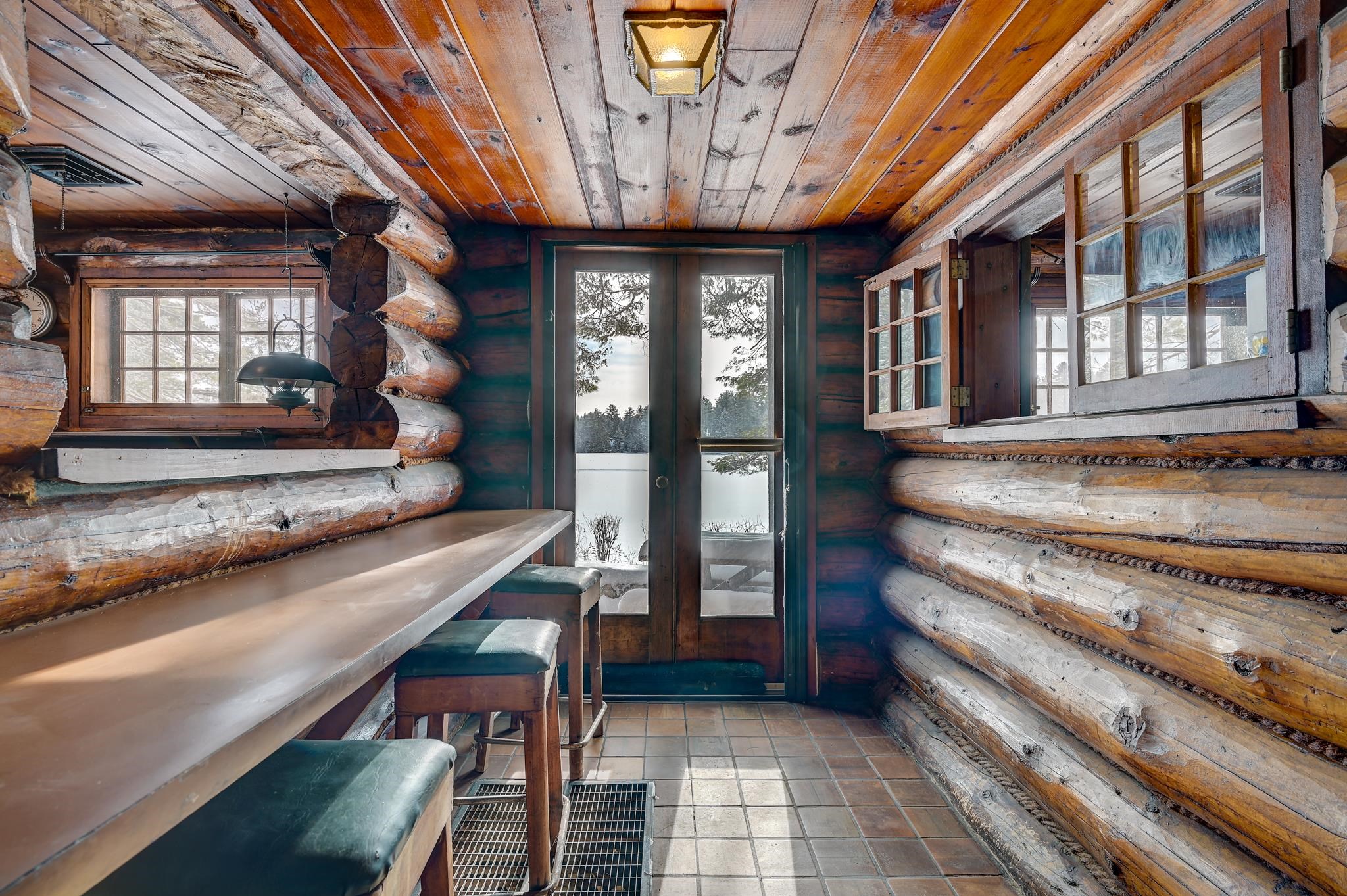 Log cabin interior with a counter and stools beside windows overlooking snow-covered trees.