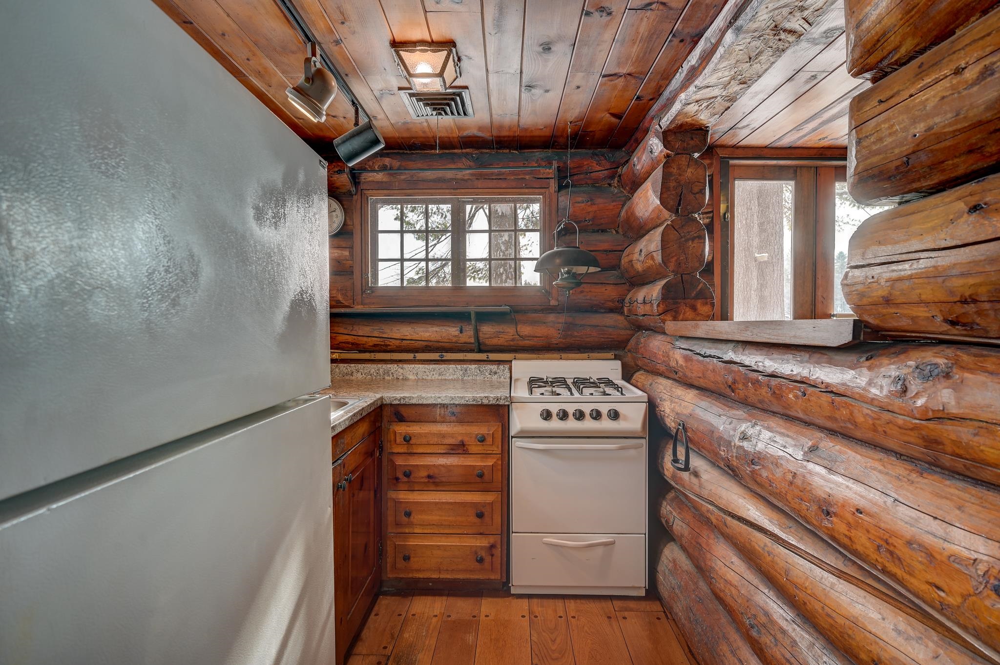 Log cabin kitchen with exposed timber walls, a white refrigerator, and a gas range beneath a window.