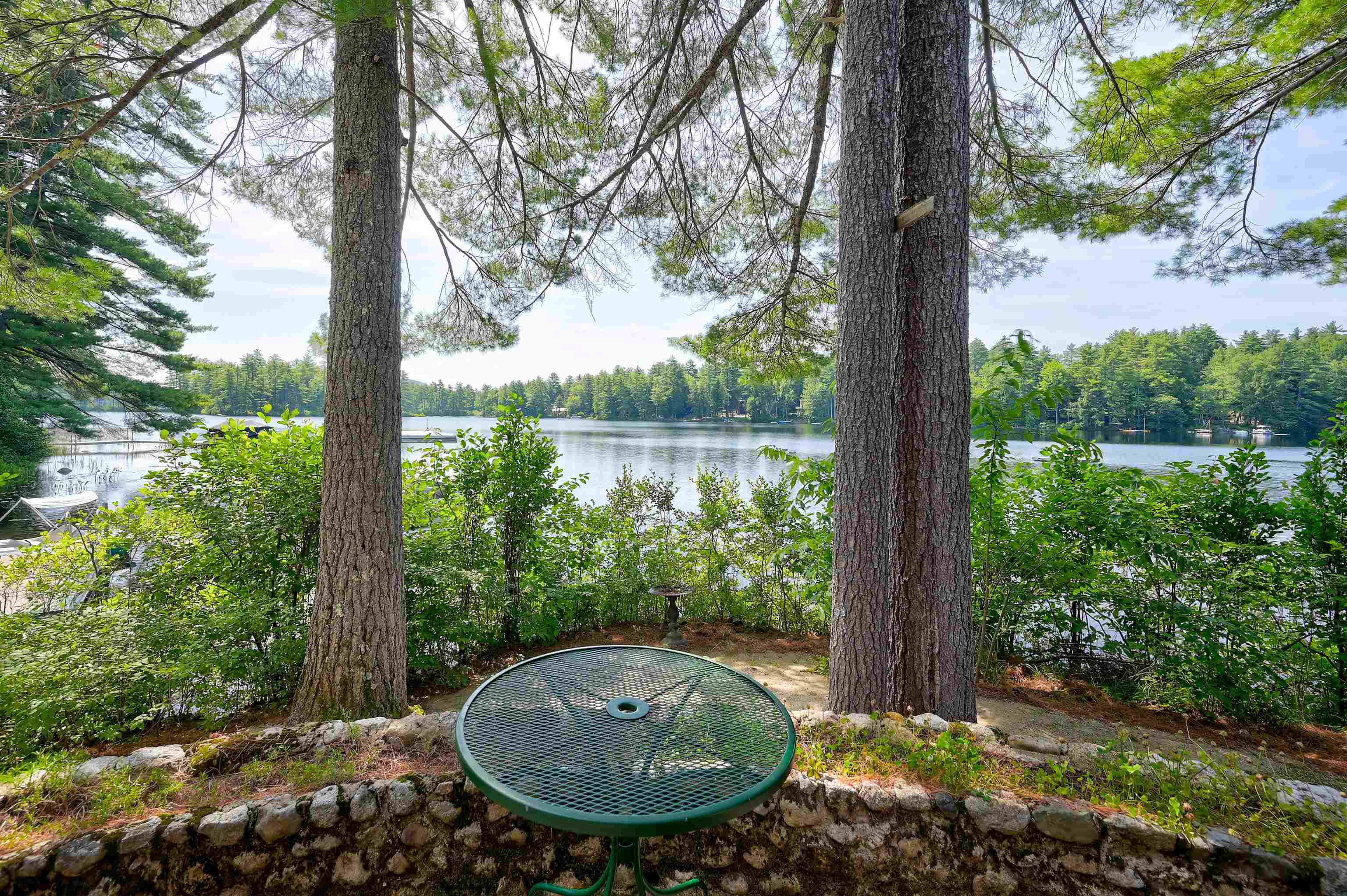 Lakeside patio with a round green table, stone wall, and pine trees framing the water.