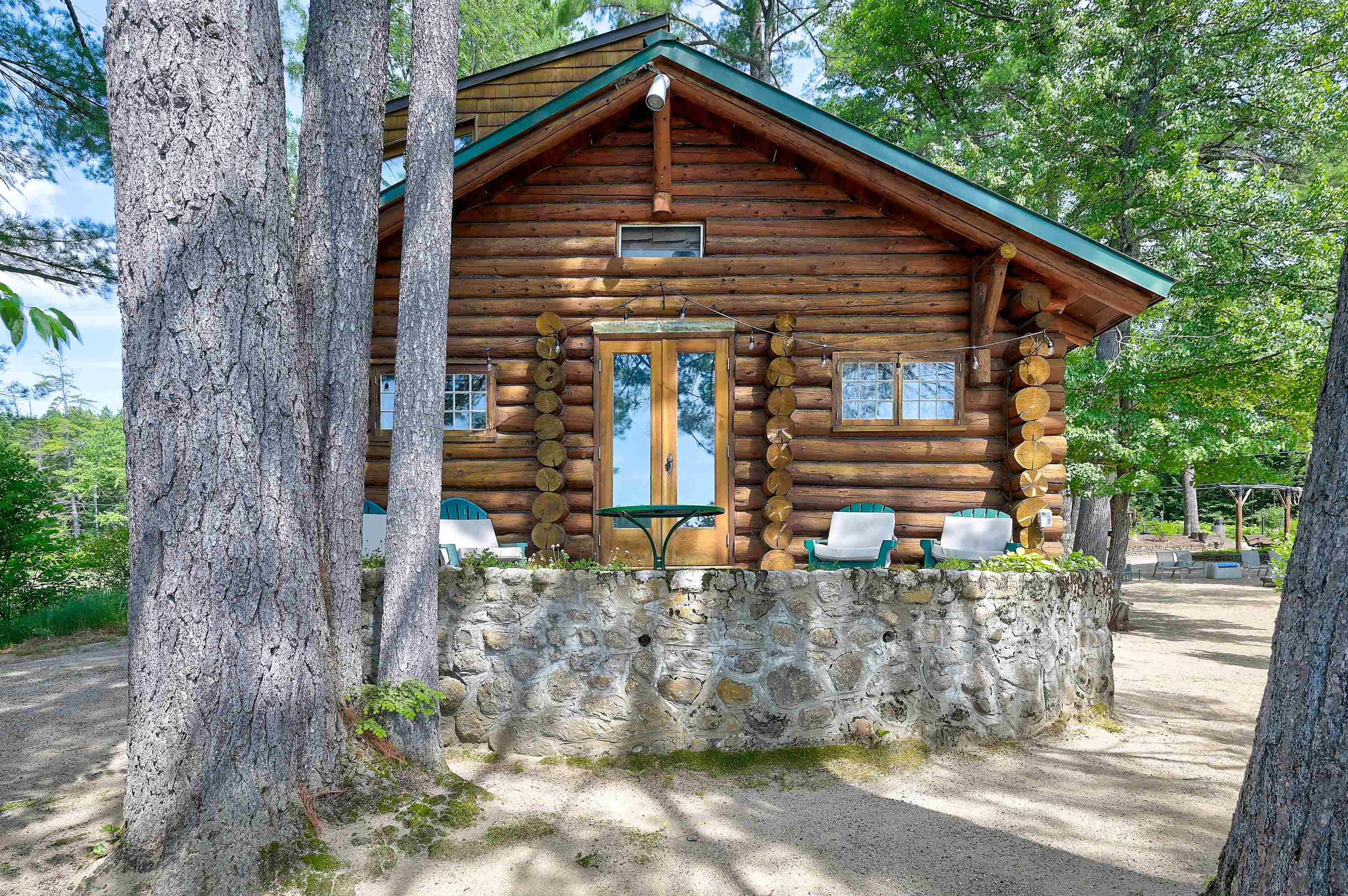 Log cabin exterior with a green roof, glass doors, outdoor chairs, and a stone wall among trees.