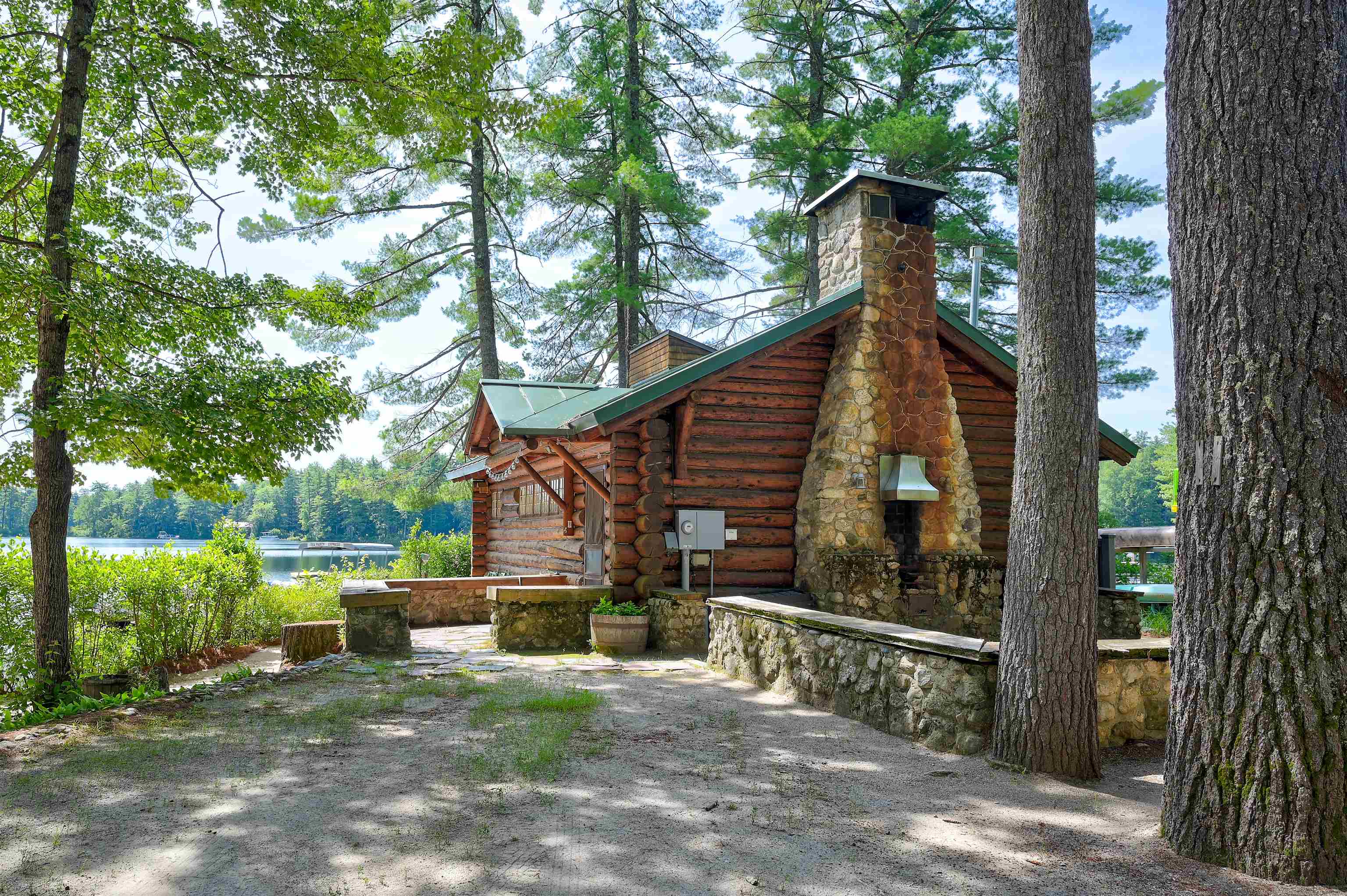 Rustic log cabin with a stone chimney and patio beside a lake, surrounded by tall trees.