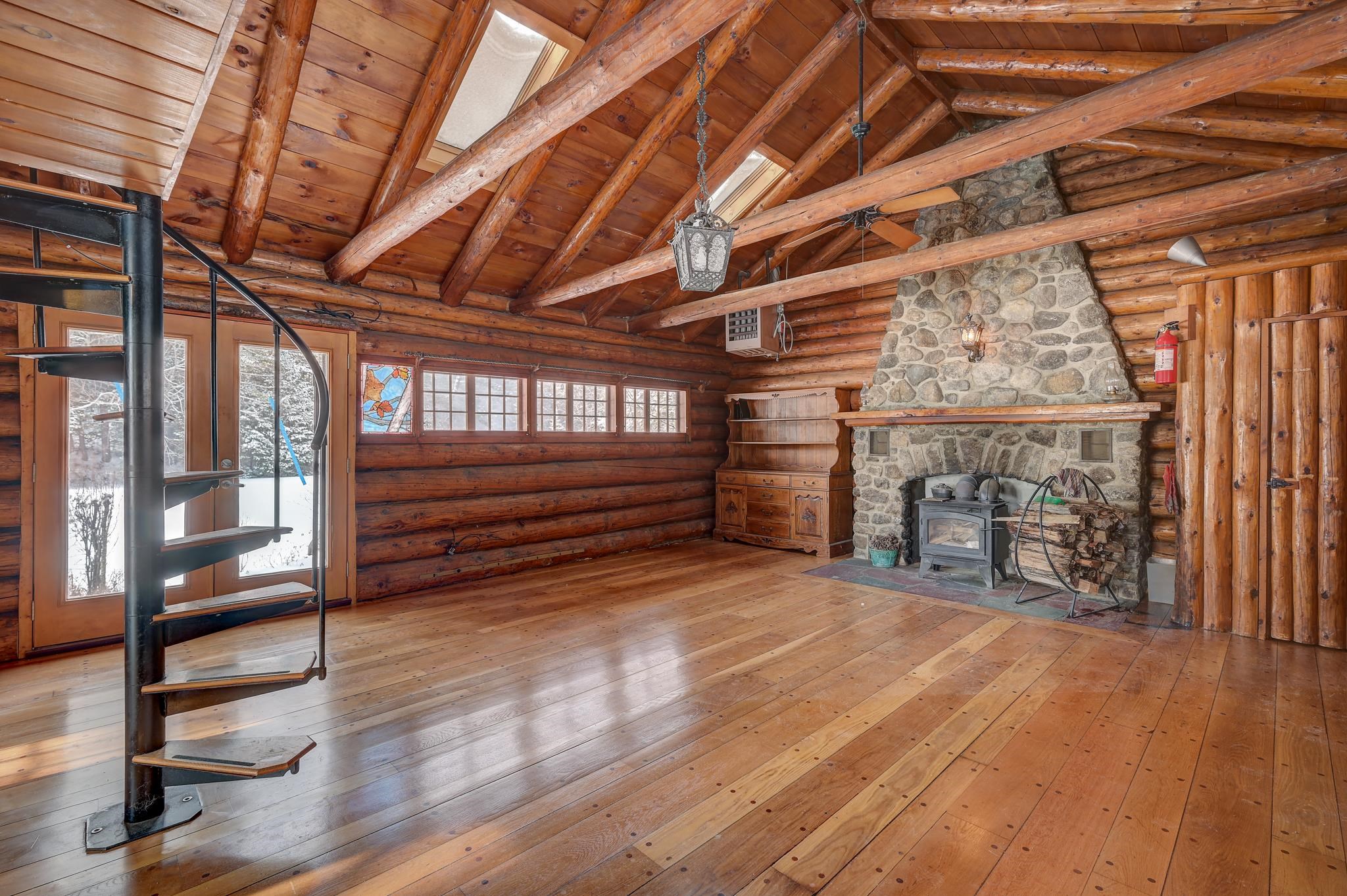 Log home living room with exposed timber rafters, spiral staircase, and large stone fireplace.