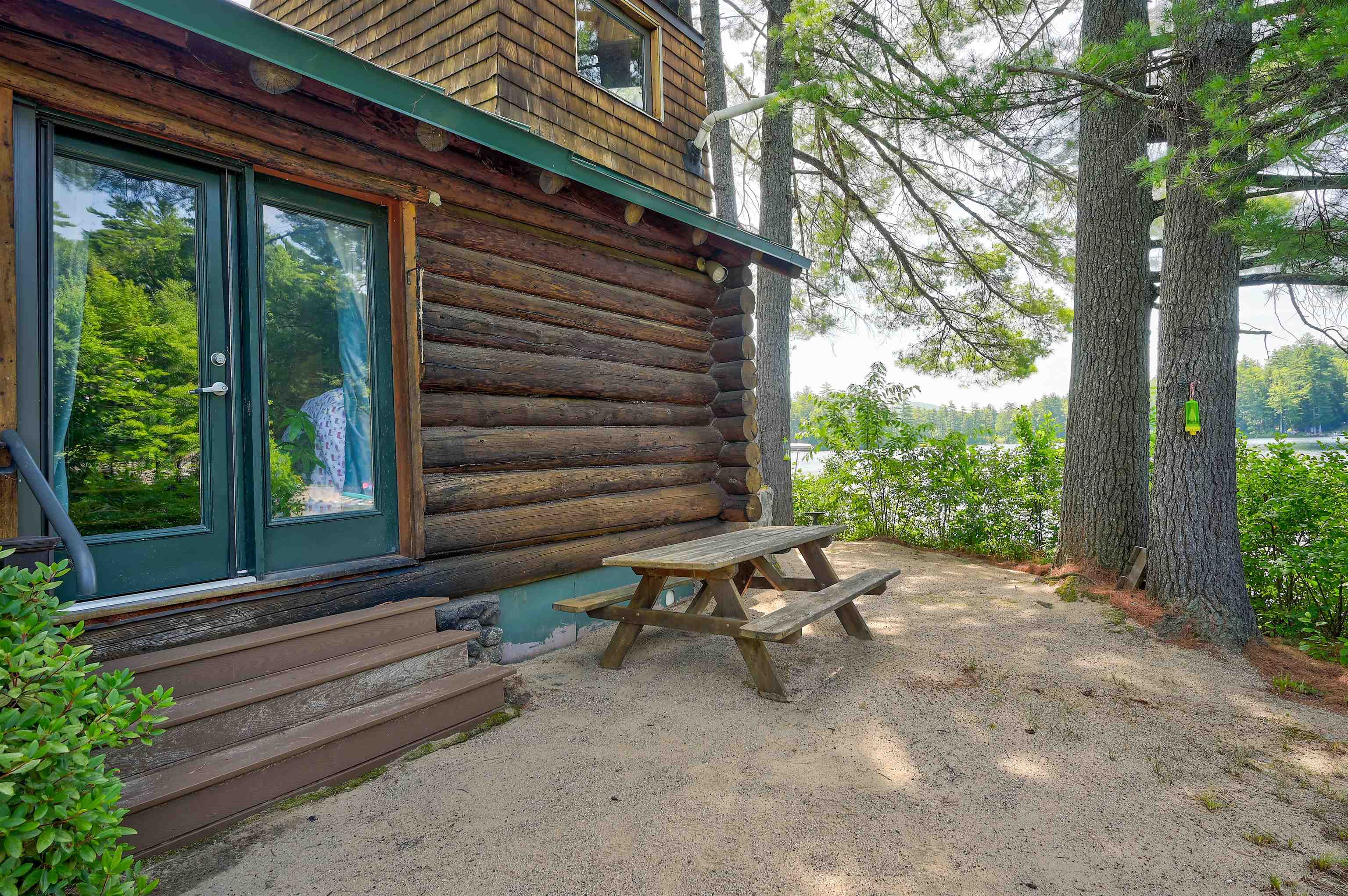 Log cabin patio with a wooden picnic table beneath trees beside a lake.