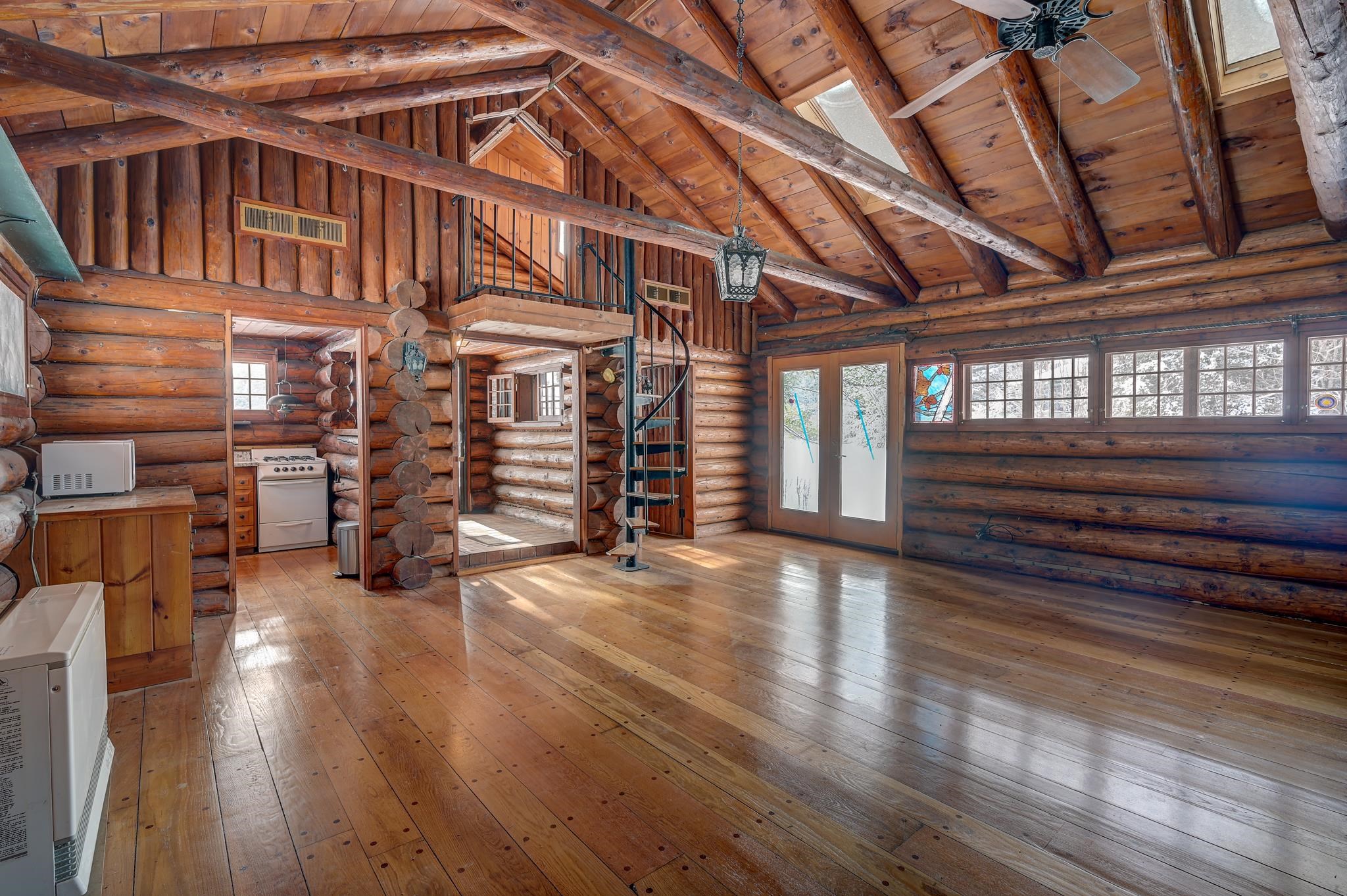 Vacant log cabin great room with exposed timber rafters, spiral staircase, hardwood floors, and a wall of windows.