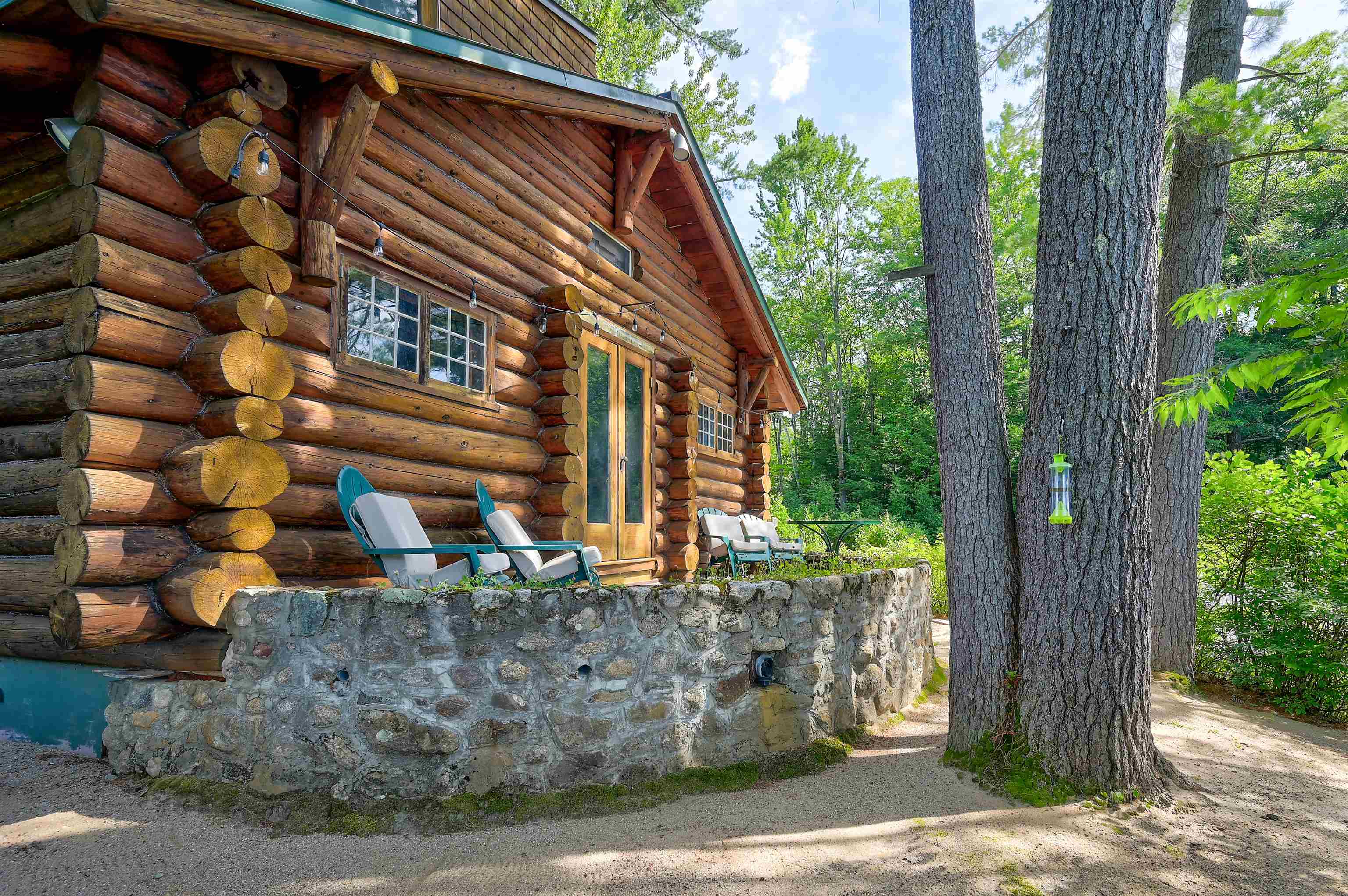 Rustic log cabin with a stone terrace wall, Adirondack chairs, and tall trees.