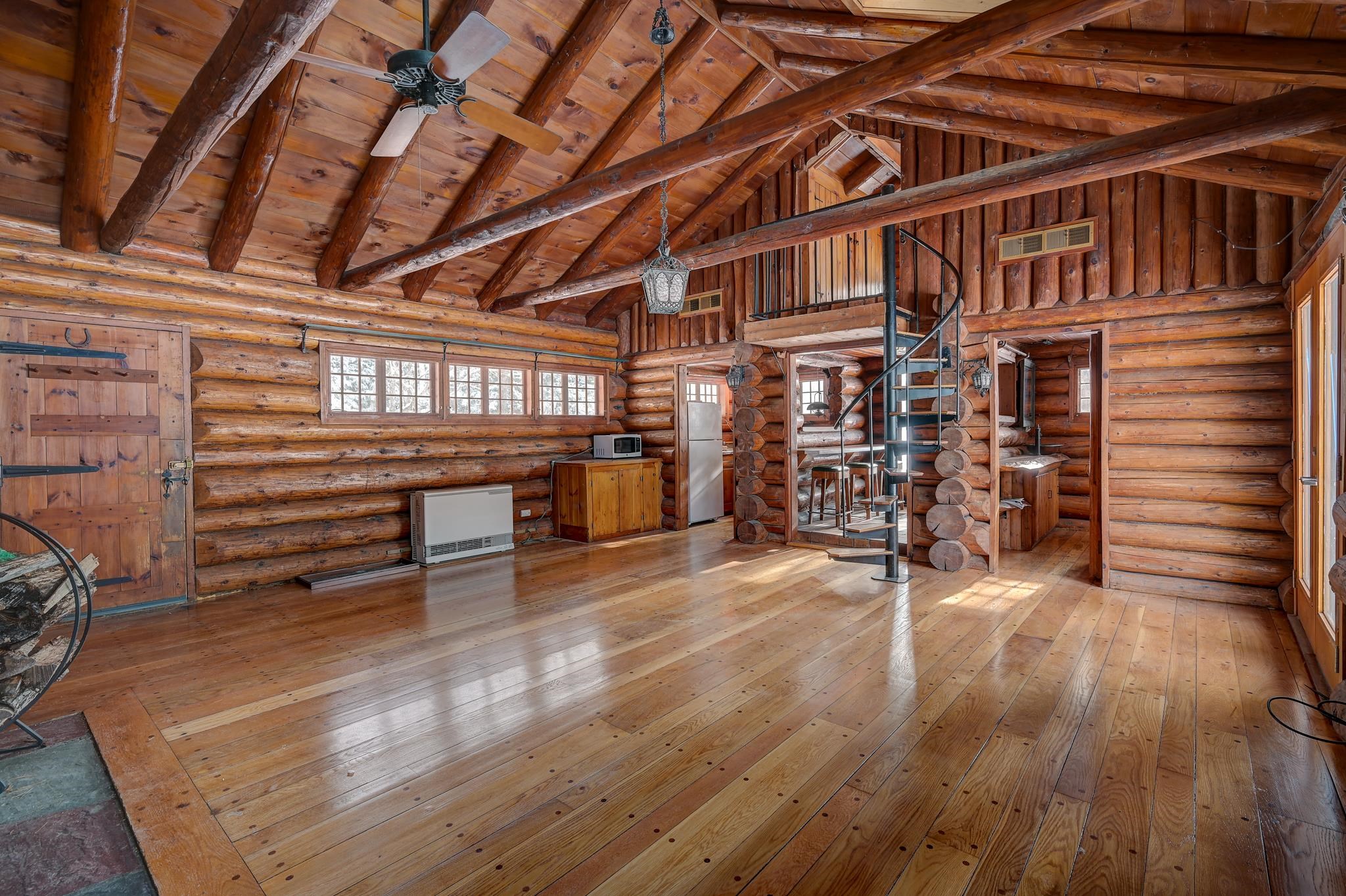 Log great room with exposed timber beams, wide plank floors, and a black spiral staircase to a loft.